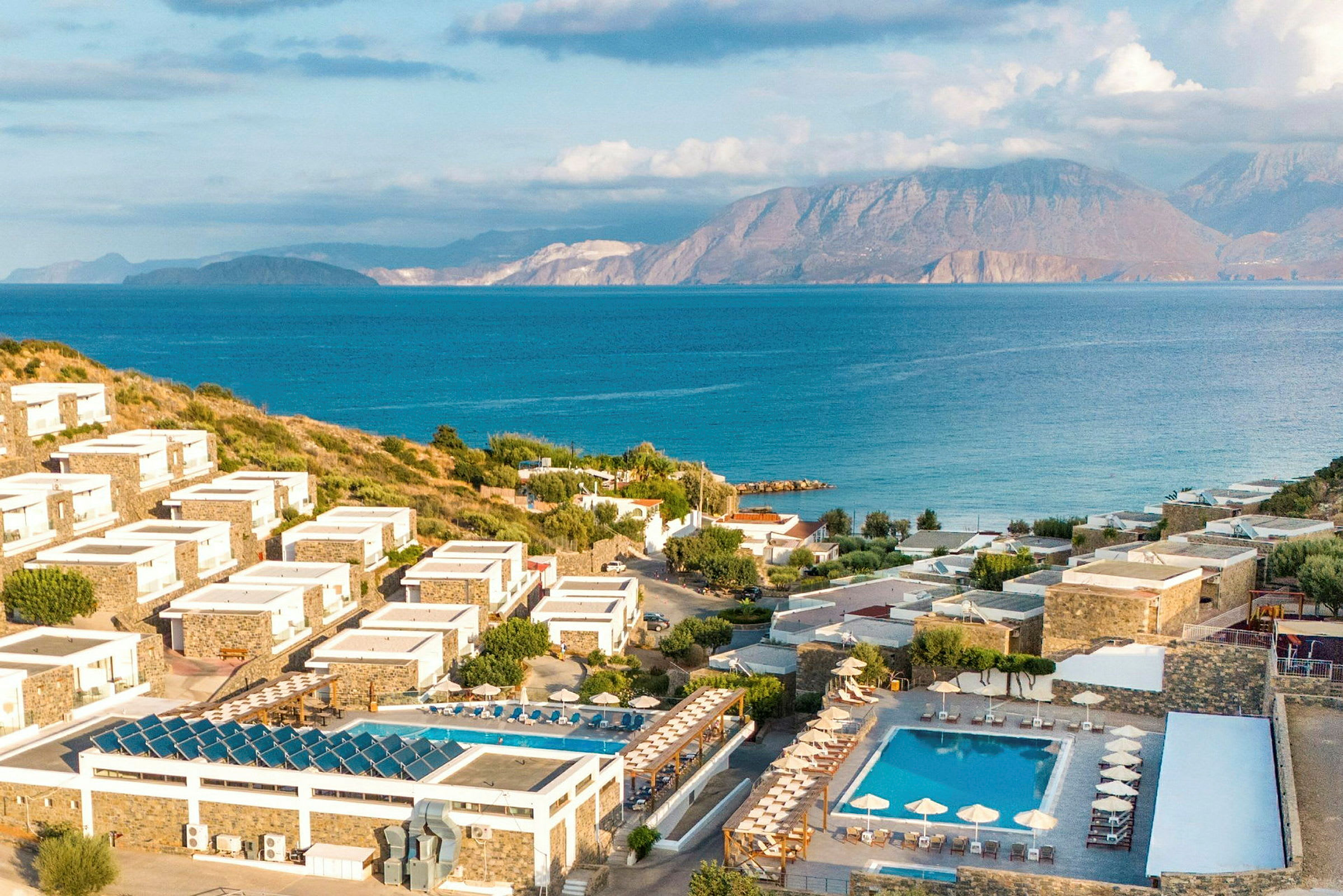 Bilde av et kystnært feriested med hotellbygninger på skrånende terreng, omgitt av grønne områder og bassenger, med hav og fjell i bakgrunnen. (AI-generert tekst)