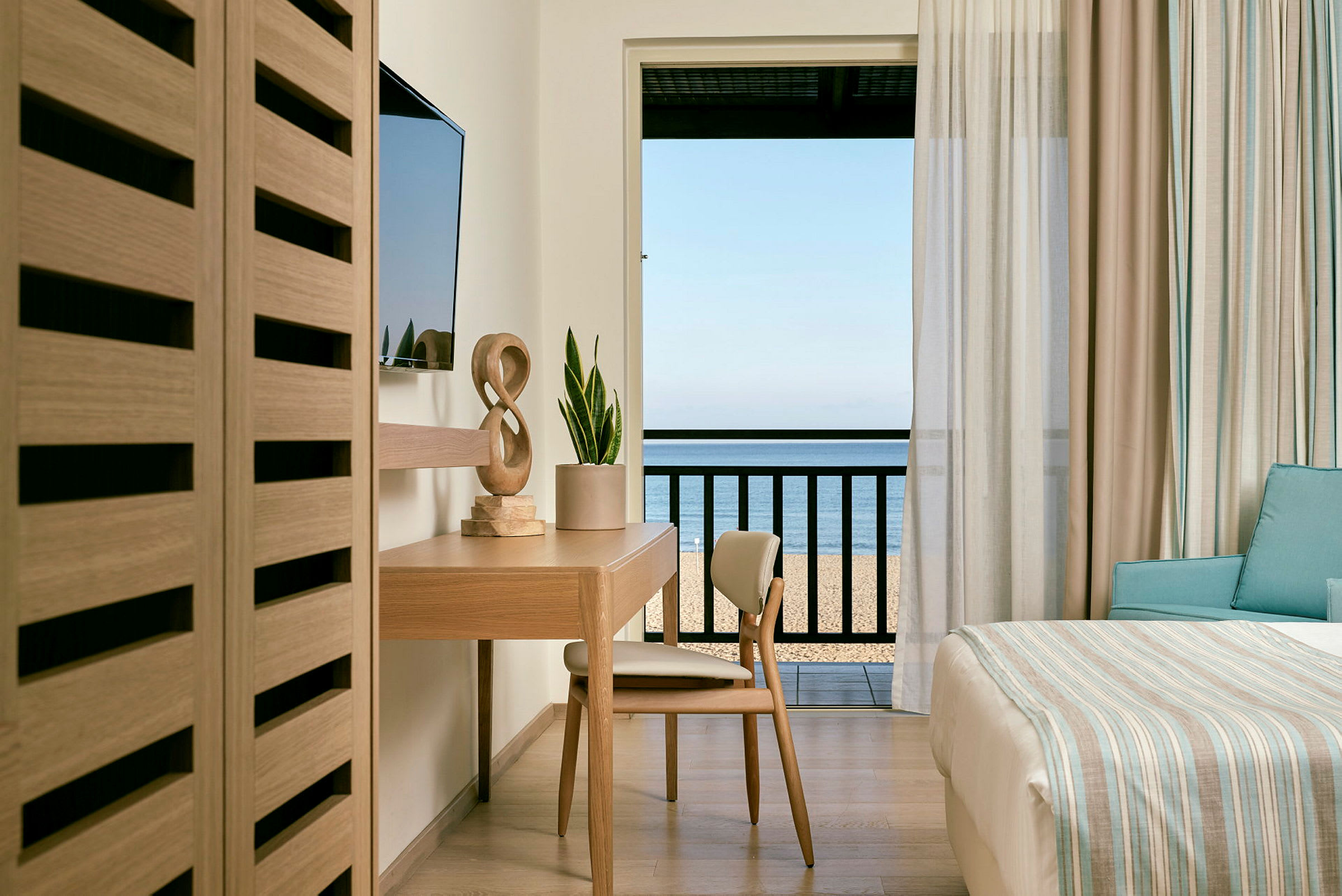 Hotellrom med skrivebord, stol og seng med utsikt mot havet fra balkongen. (AI-generert tekst)
