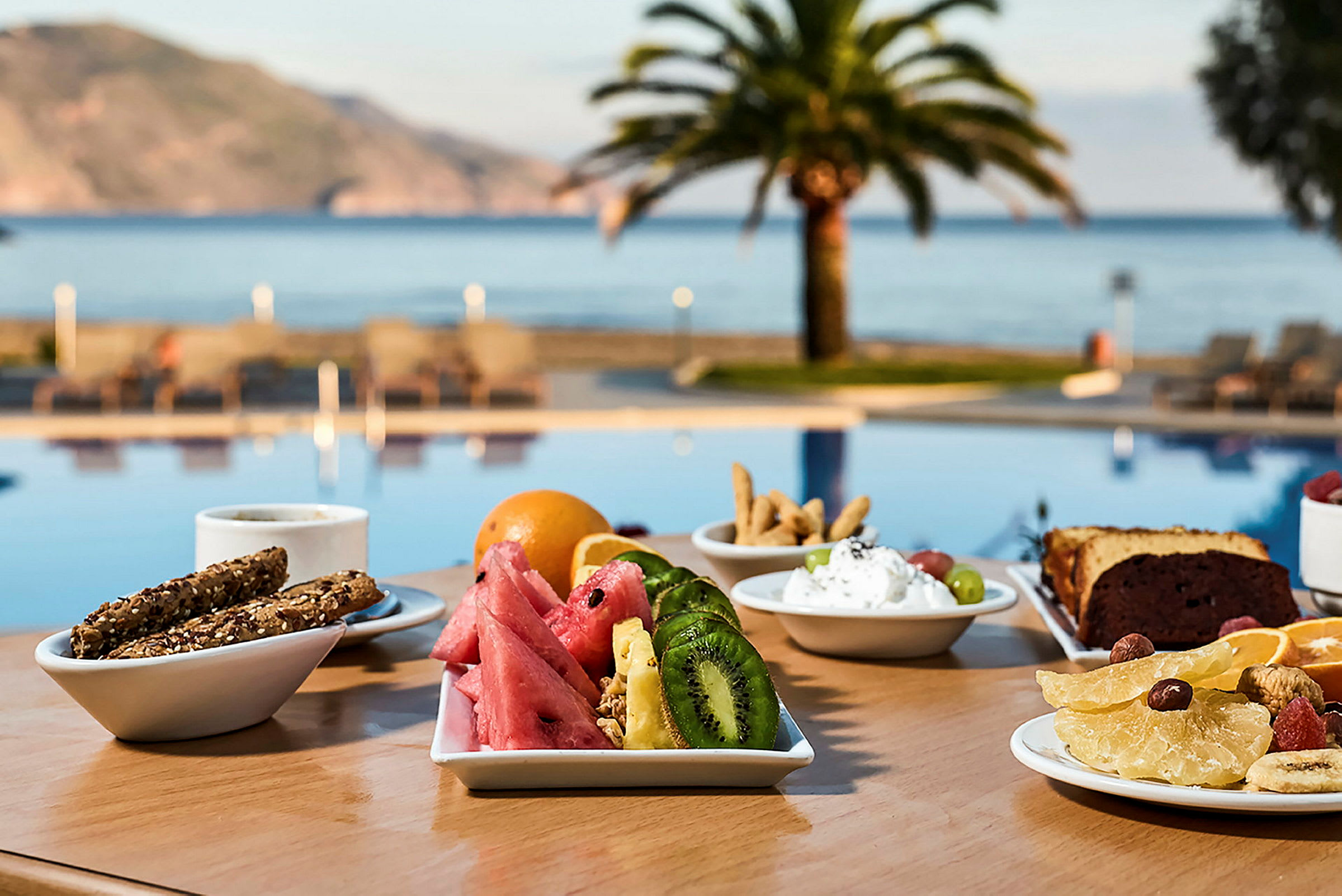 En frokost serveres på et bord nær et basseng, med utsikt mot havet og palmer. (AI-generert tekst)