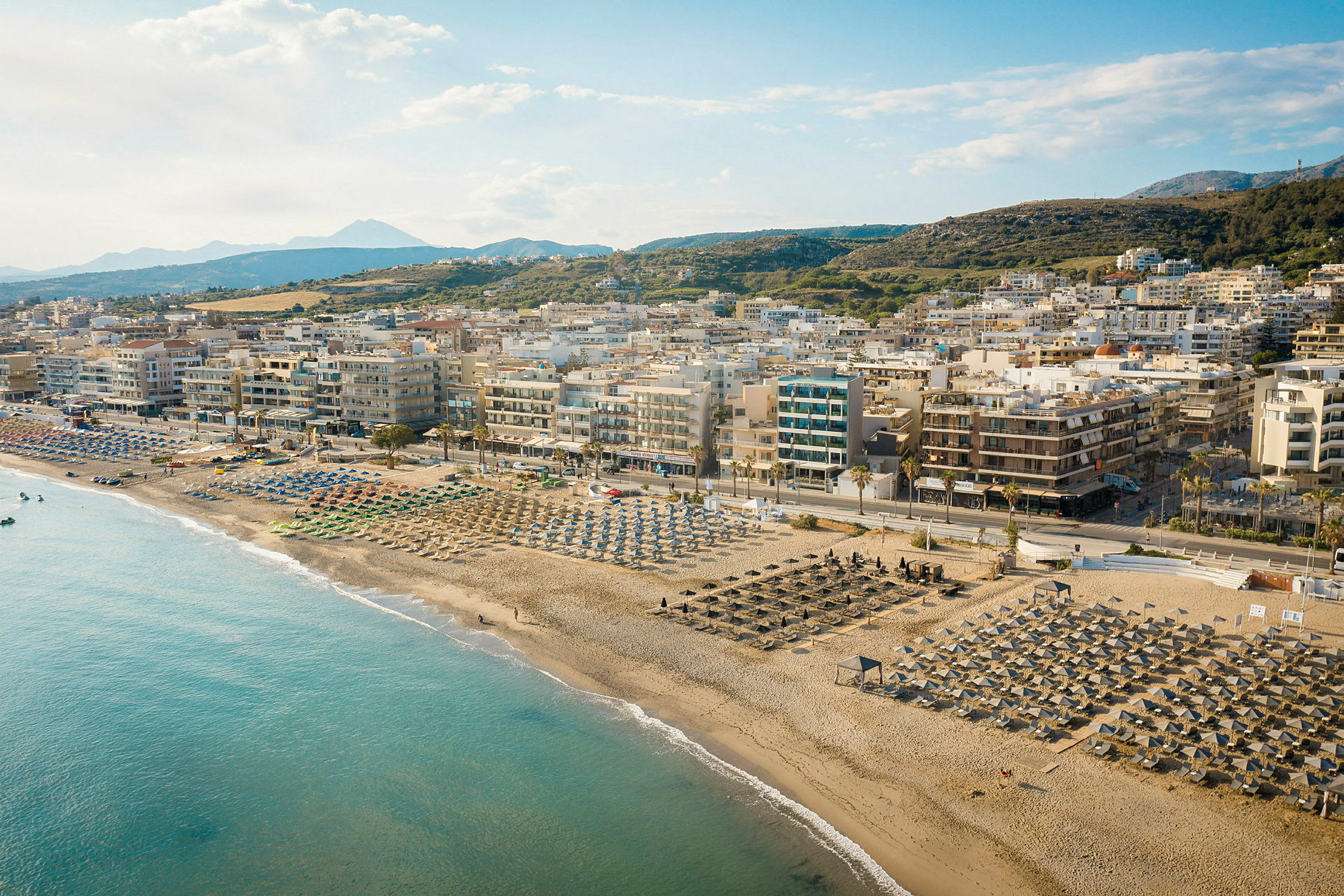 En sandstrand med parasoller og solsenger foran en by med leilighetsbygg og fjell i bakgrunnen. (AI-generert tekst)