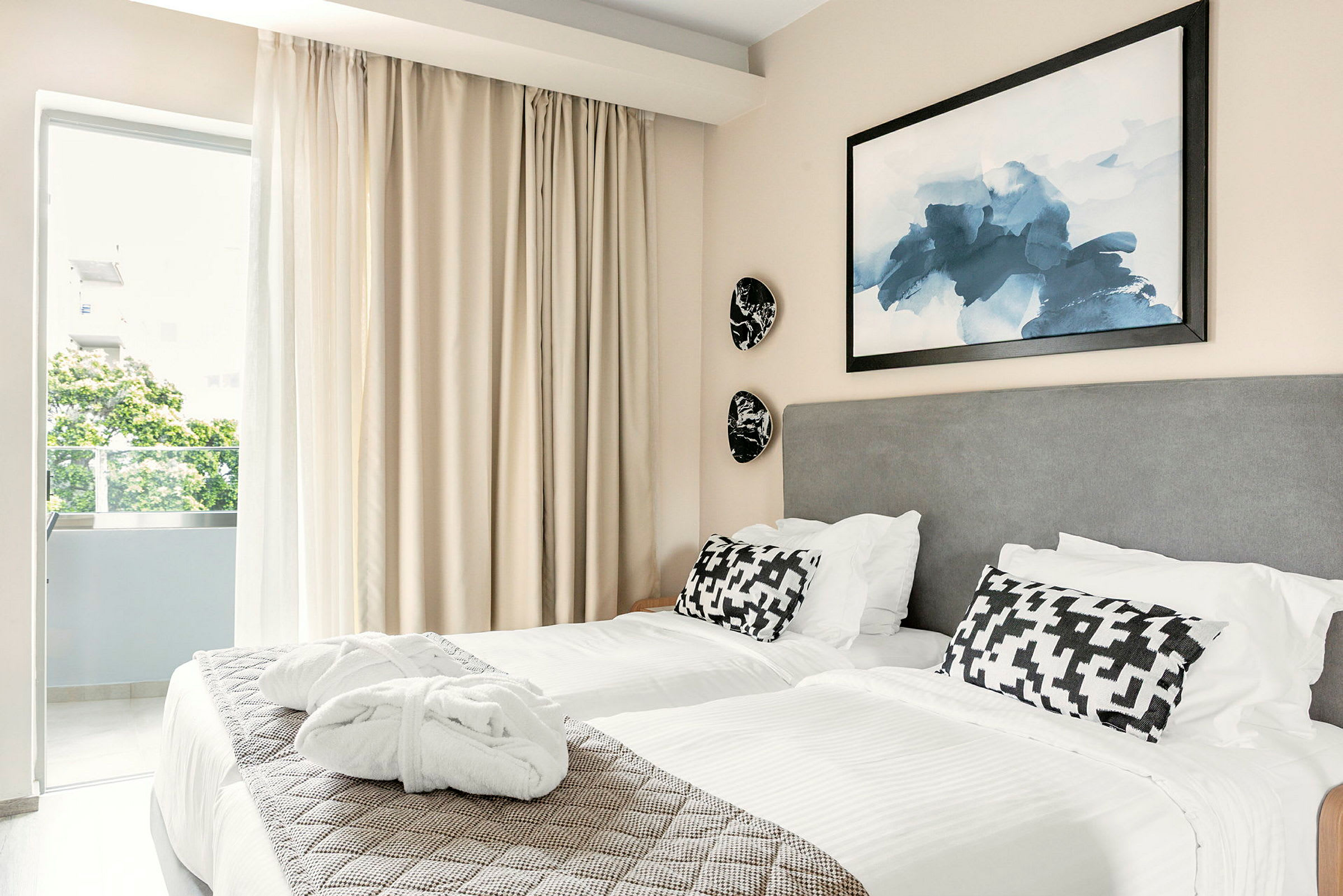 To senger med hvite laken og dekorative puter på et hotellrom. Et bilde henger over sengene, og gardiner er trukket foran vinduet. (AI-generert tekst)