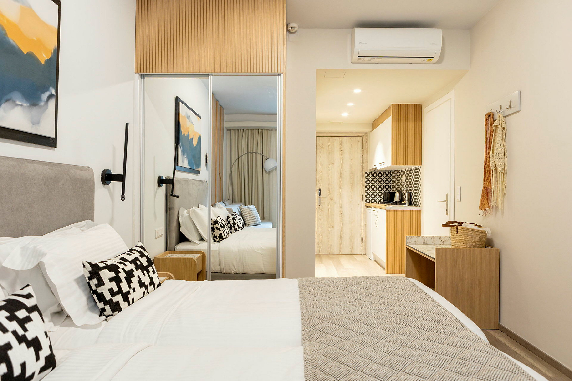 Et moderne hotellrom med en dobbeltseng, hvitt sengetøy og dekorative puter. På veggen henger et abstrakt maleri, og i bakgrunnen er det et lite kjøkken. (AI-generert tekst)