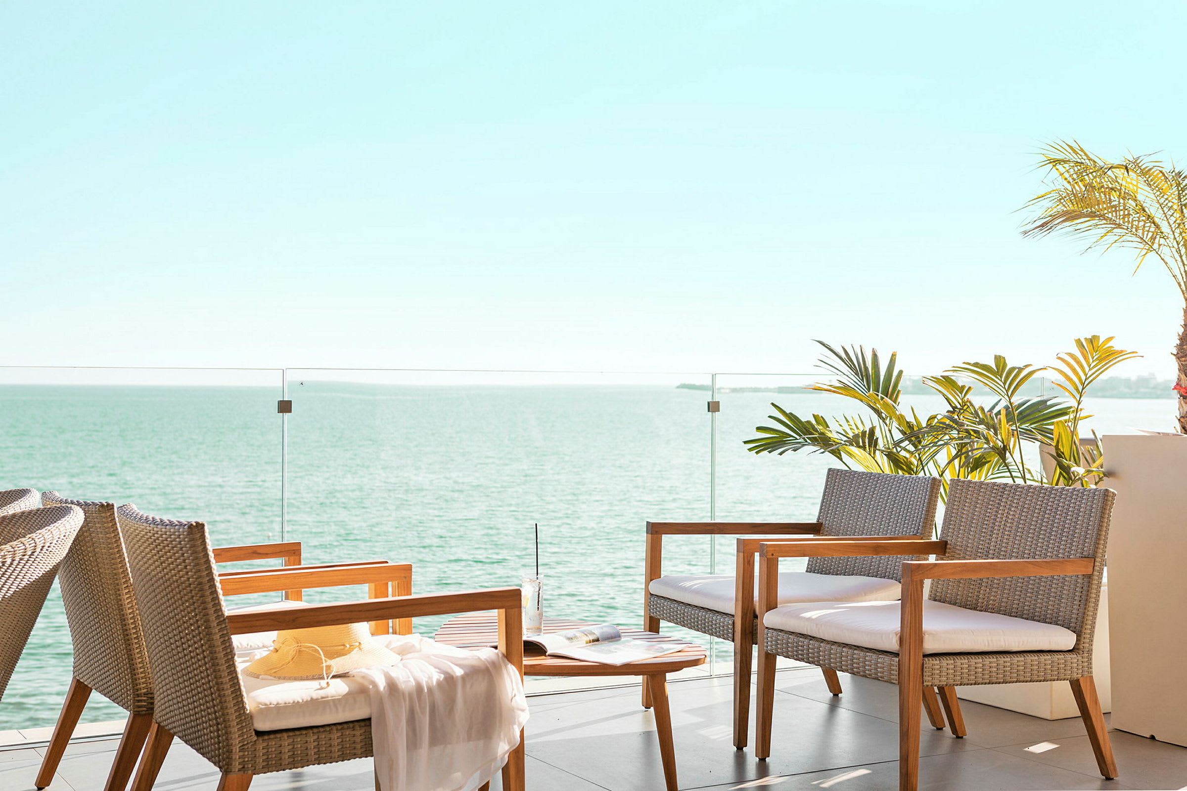 En sittegruppe med stoler og bord på en terrasse med glassrekkverk. I bakgrunnen sees havet og himmelen. (AI-generert tekst)