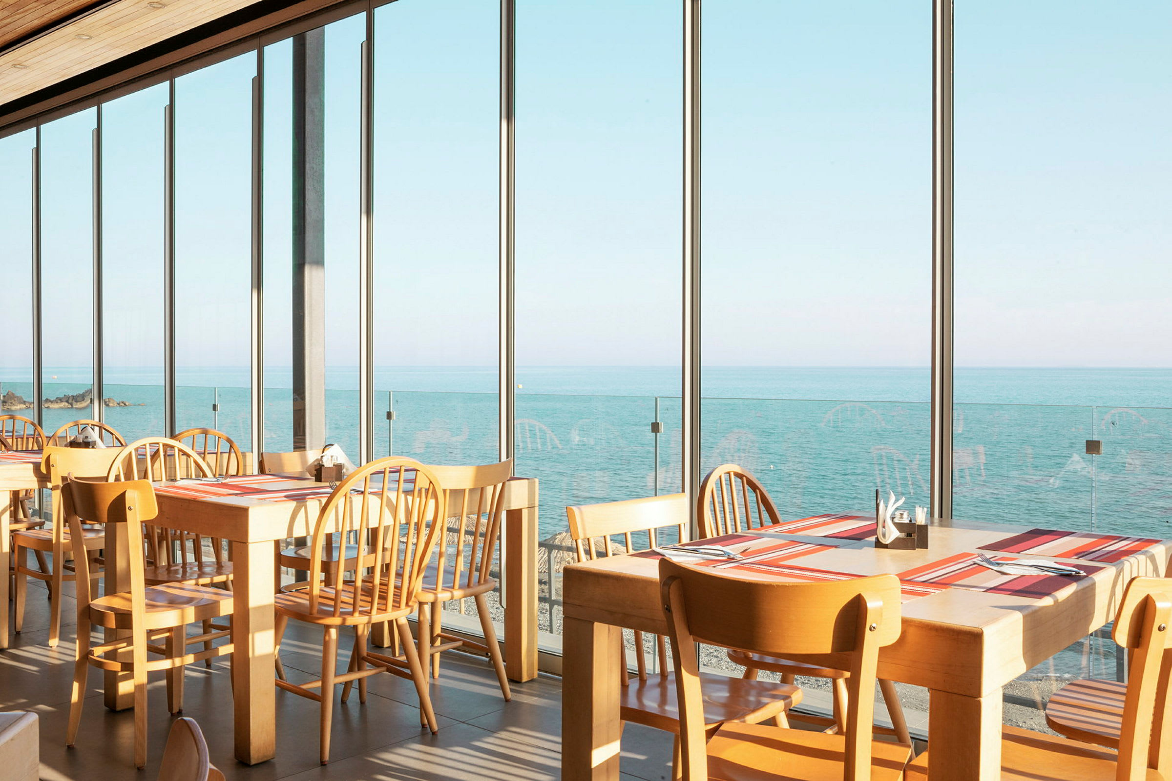 Restaurant med bord og stoler ved store vinduer som vender mot havet. (AI-generert tekst)