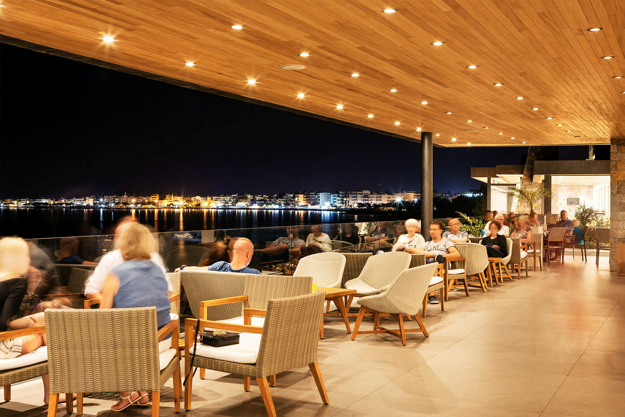 En restaurant med uteområde under en trepanelert takterrasse, med utsikt over en opplyst by ved vannet om kvelden. (AI-generert tekst)