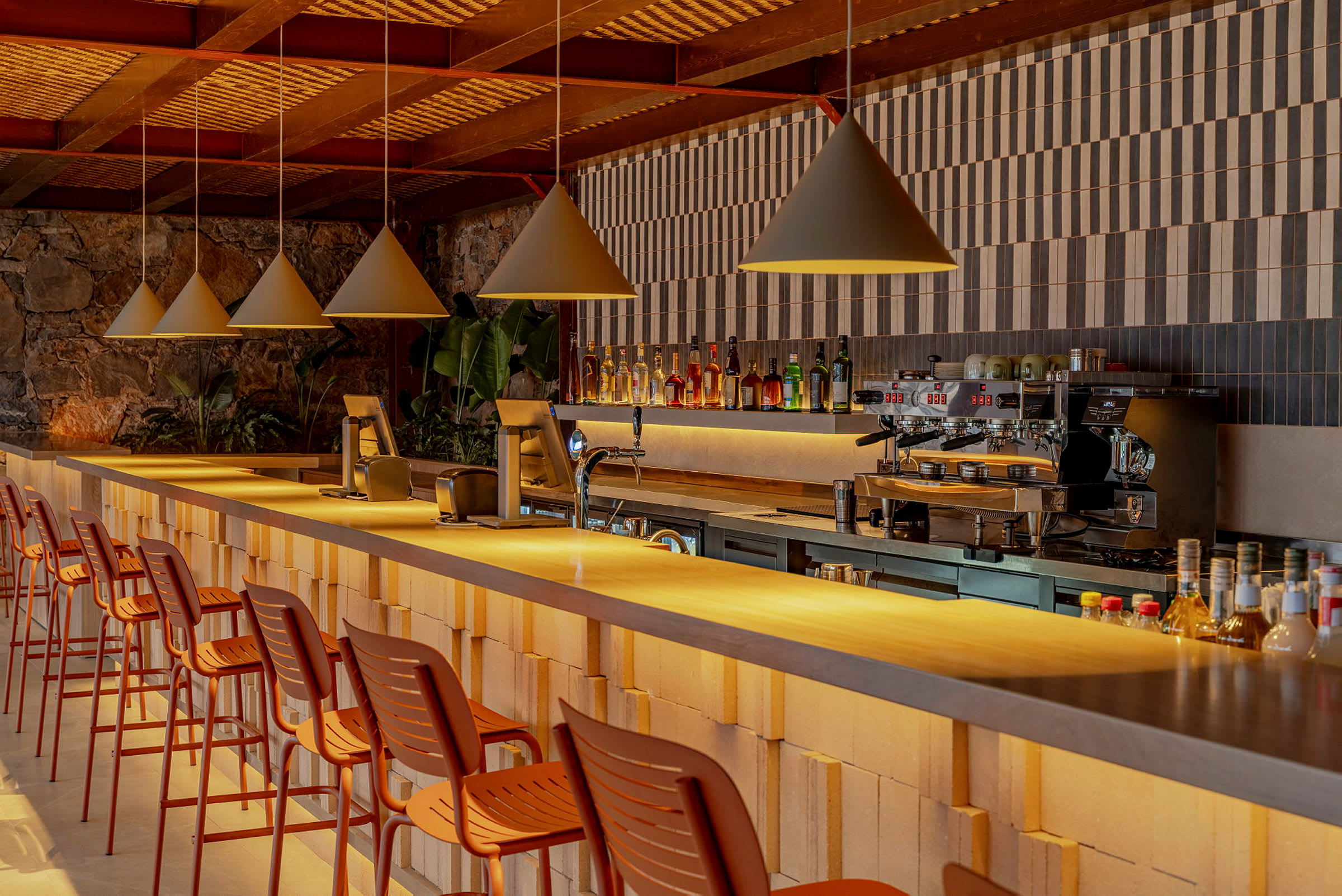 Bildet viser en moderne bar med en rekke høye stoler ved en lang bardisk. Baren er dekorert med flasker og har en kaffemaskin. (AI-generert tekst)