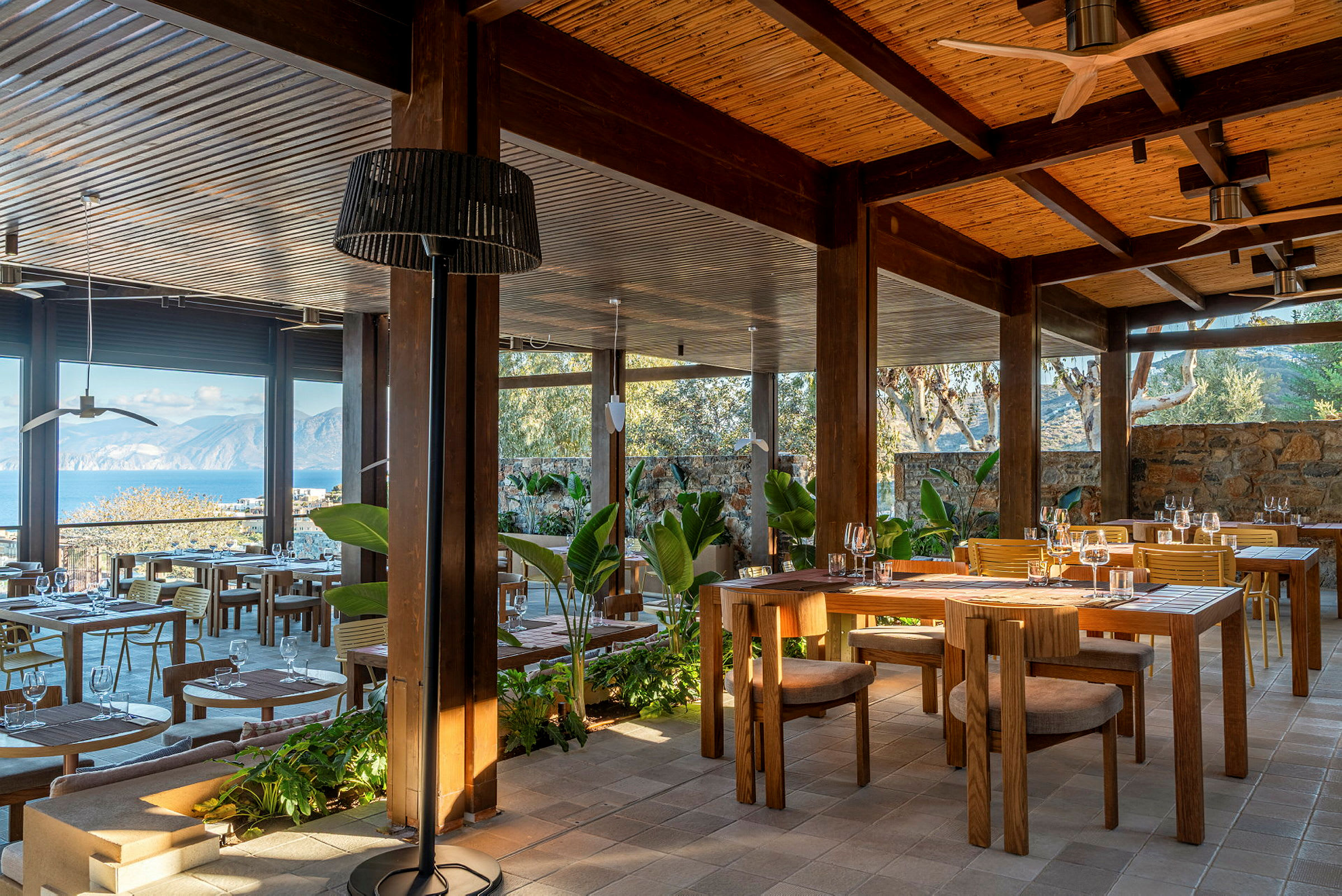 En utendørs restaurant med trebord og utsikt over havet og fjellene. (AI-generert tekst)