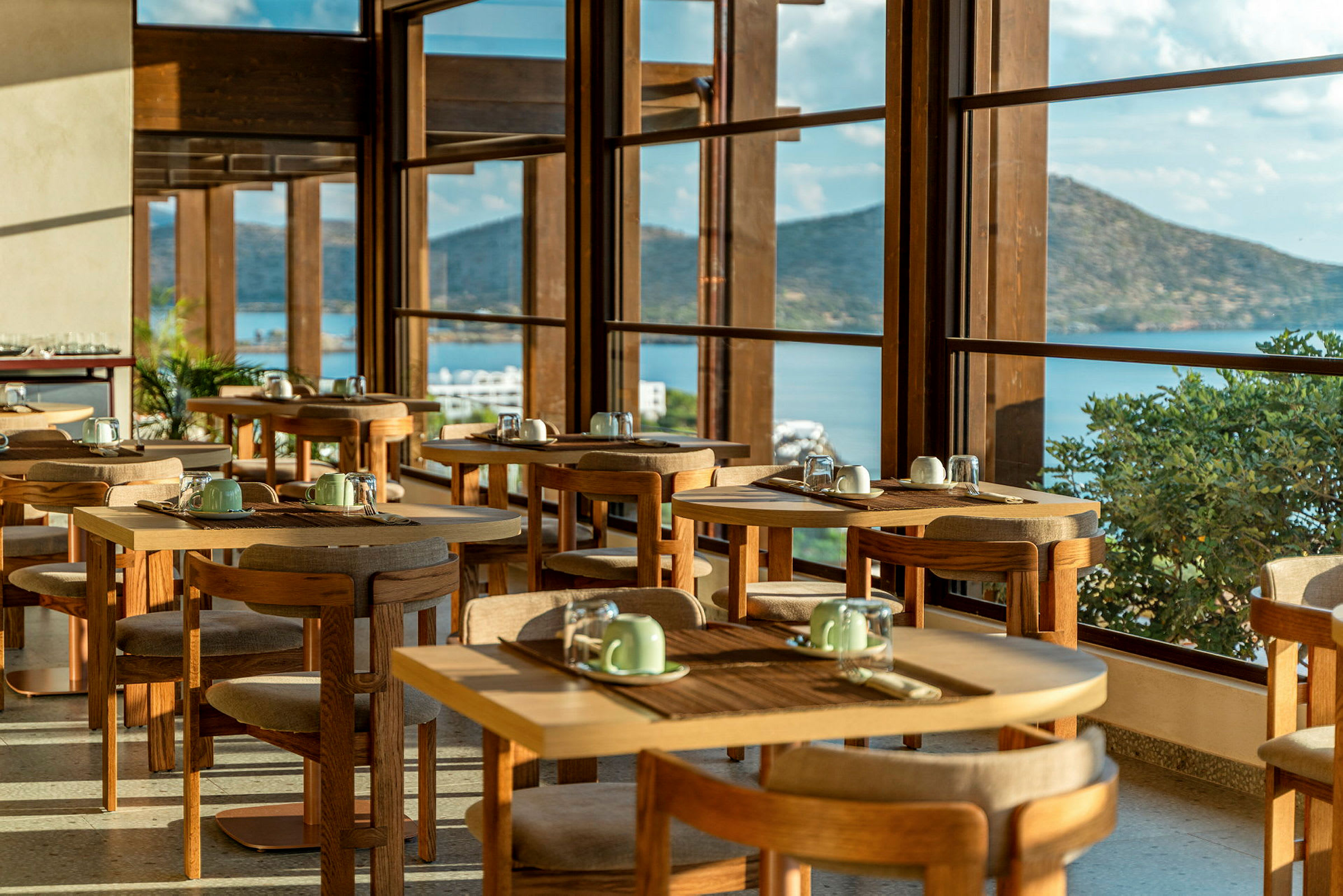 Et restaurantområde med bord og stoler, med store vinduer som viser utsikt over en havbukt og fjell. (AI-generert tekst)