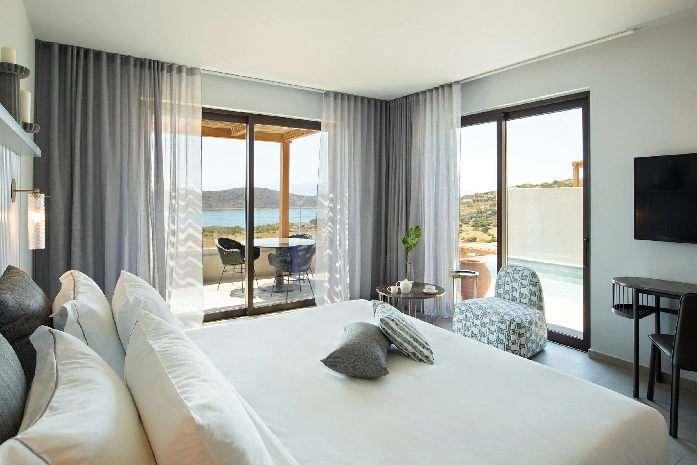Moderne hotellrom med stor seng, sittegruppe og utsikt mot sjø og landskap gjennom glassdører. (AI-generert tekst)
