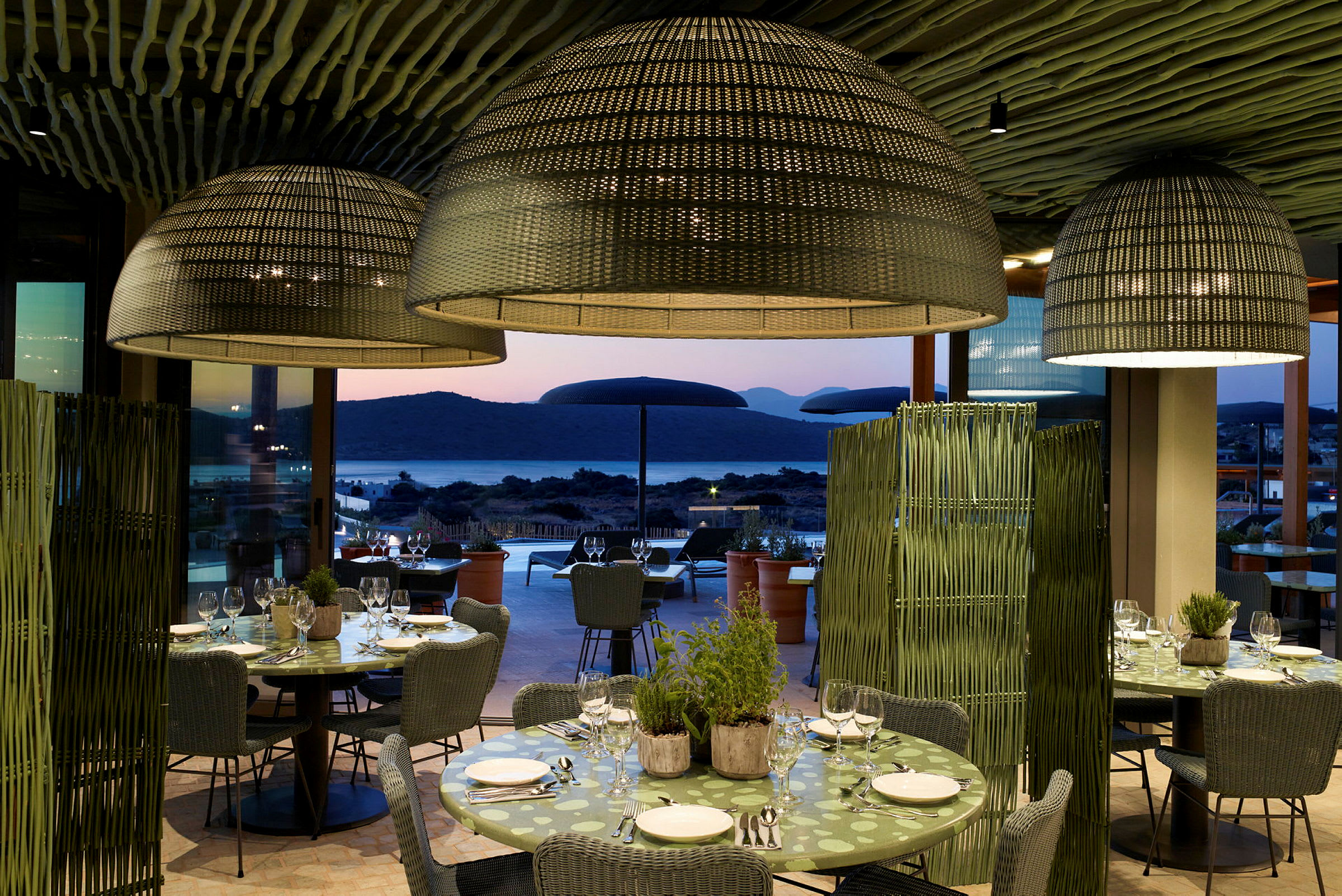 Restaurant med dekkede bord og store lampeskjermer, utsikt mot havet i solnedgang. (AI-generert tekst)
