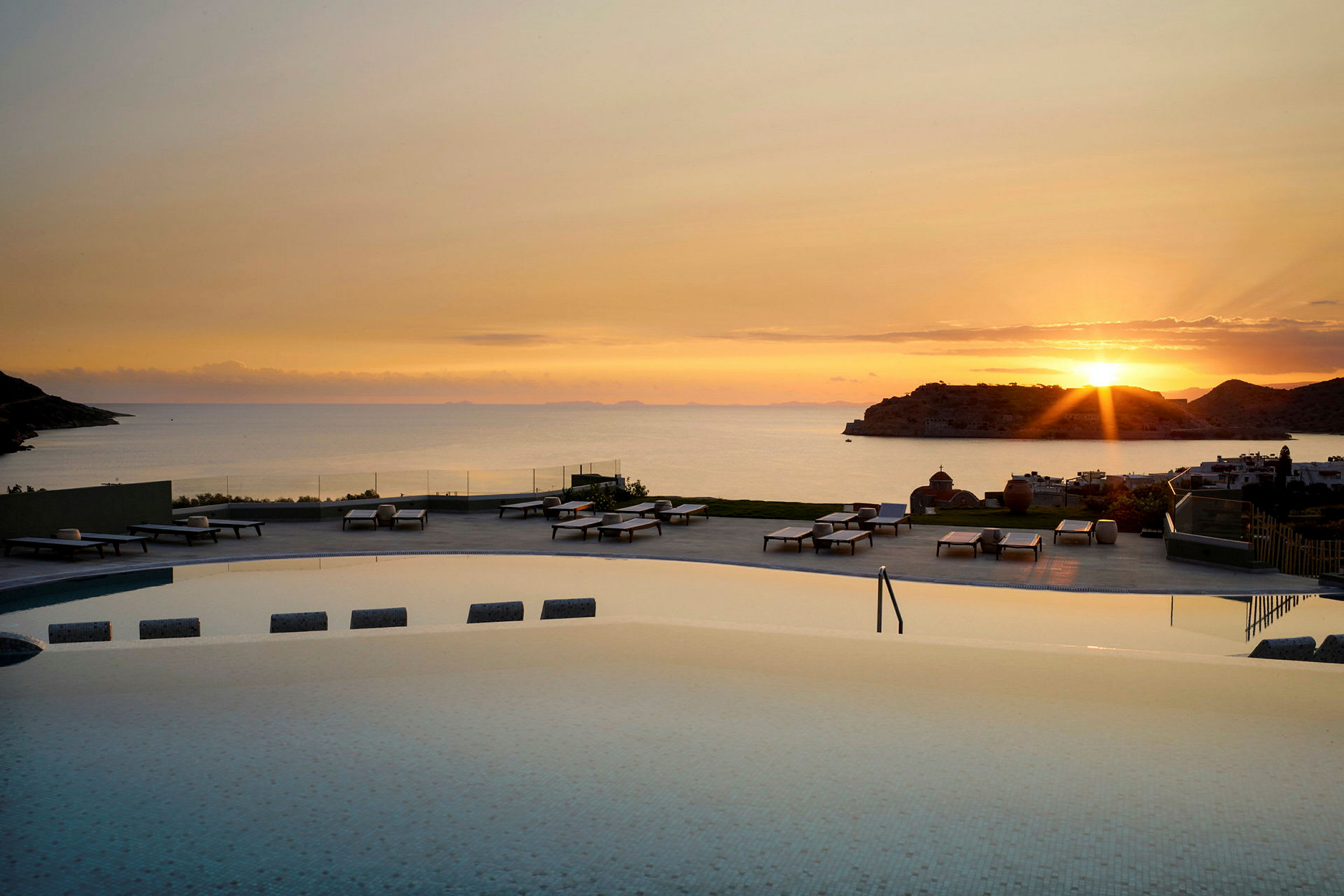 Utsikt over et basseng med solsenger i forgrunnen og havet ved solnedgang i bakgrunnen. (AI-generert tekst)