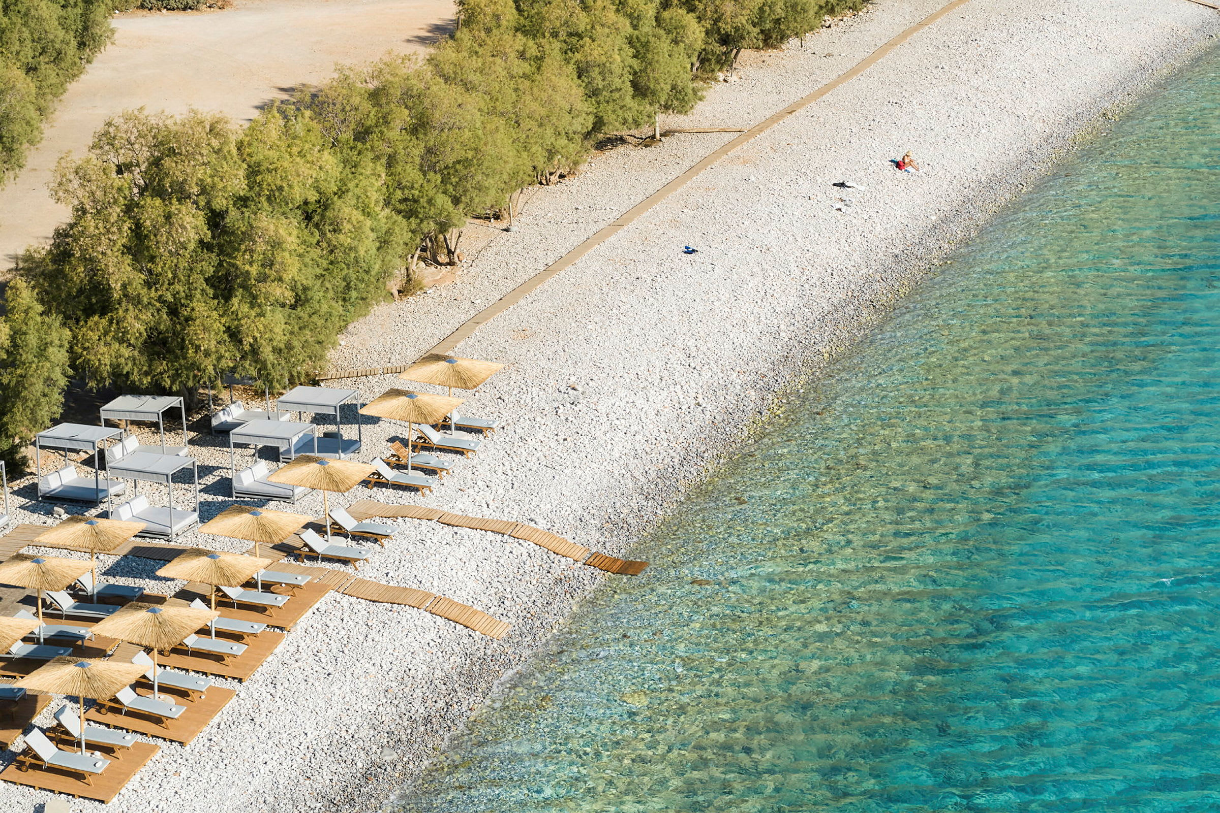 En steinete strand med solsenger, parasoller og trestier nært klart vann. (AI-generert tekst)