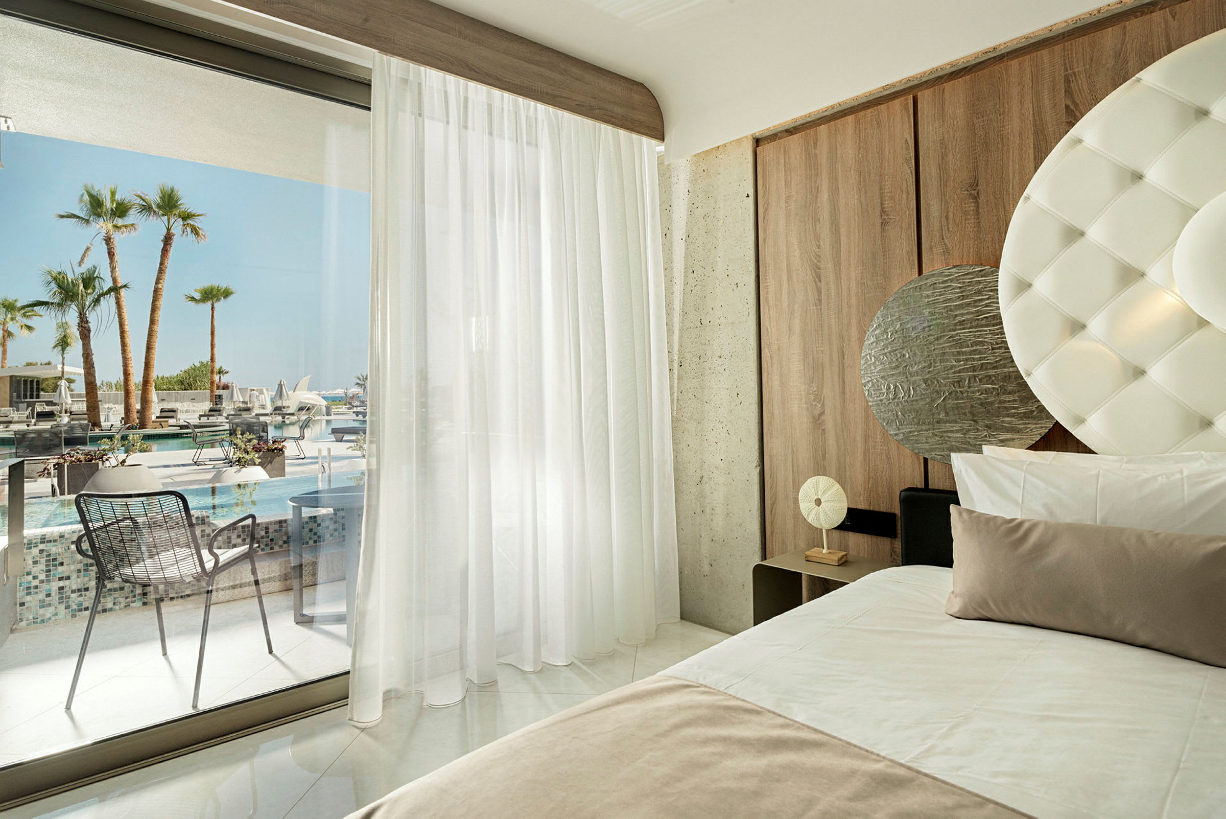 Moderne hotellrom med jordtoner og utsikt mot bassengområdet og palmene gjennom glassdører. (AI-generert tekst)
