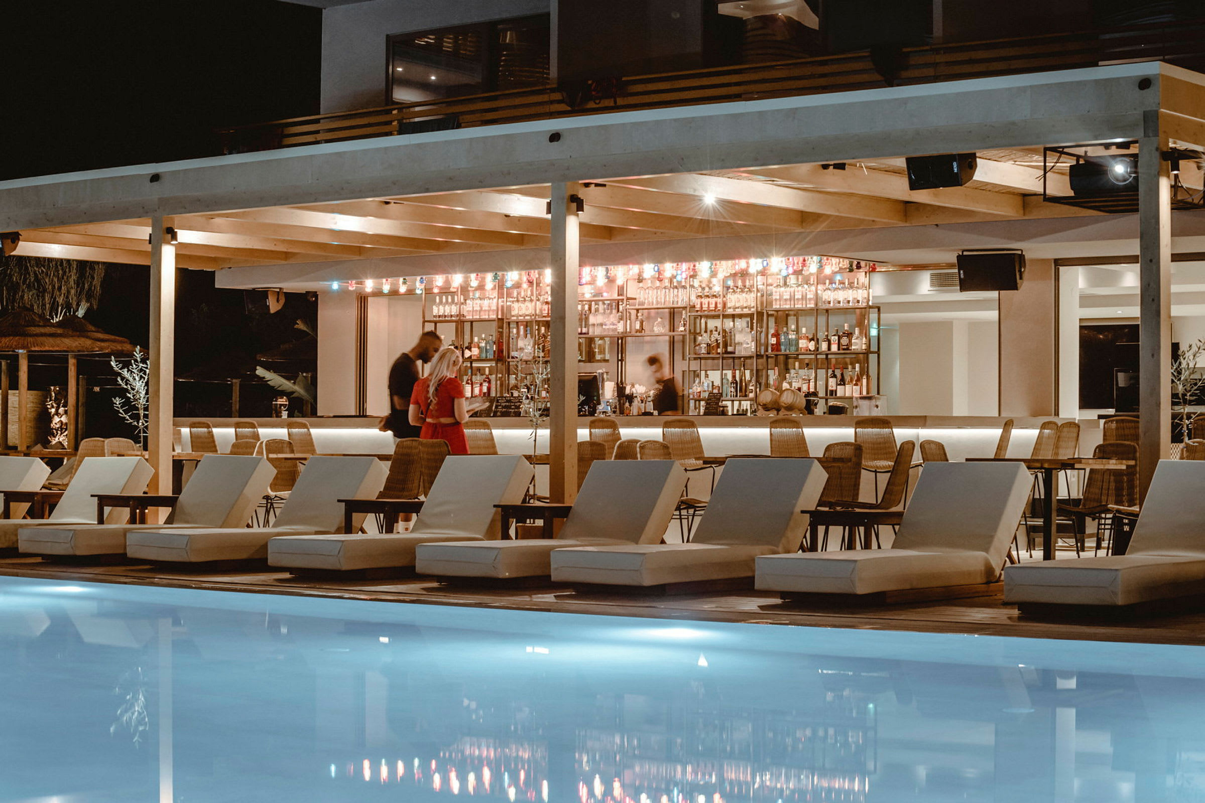 En kveldsstemning ved et luksuriøst basseng med solsenger foran en opplyst bar. (AI-generert tekst)