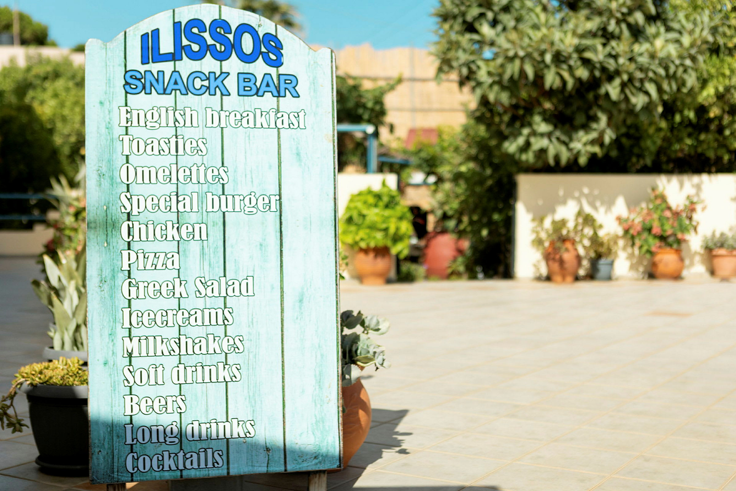 Skilt som viser menyen på Ilissos Snack Bar med ulike retter og drikkevarer. (AI-generert tekst)