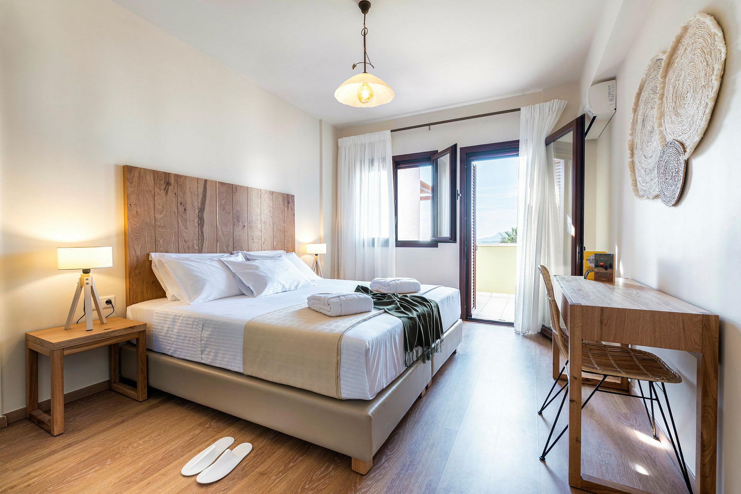 Et lyst og innbydende hotellrom med en dobbeltseng, to nattbord og en skrivepult. Vinduer står åpne mot en balkong med utsikt. (AI-generert tekst)