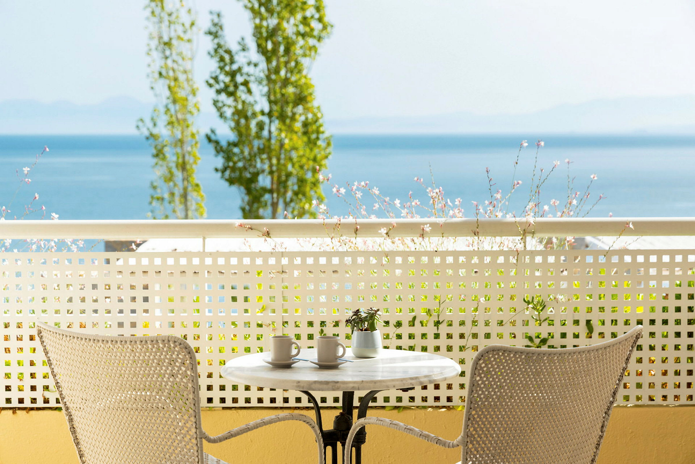 Utsikt fra en terrasse med et bord og to stoler, blomster og hav i bakgrunnen. (AI-generert tekst)