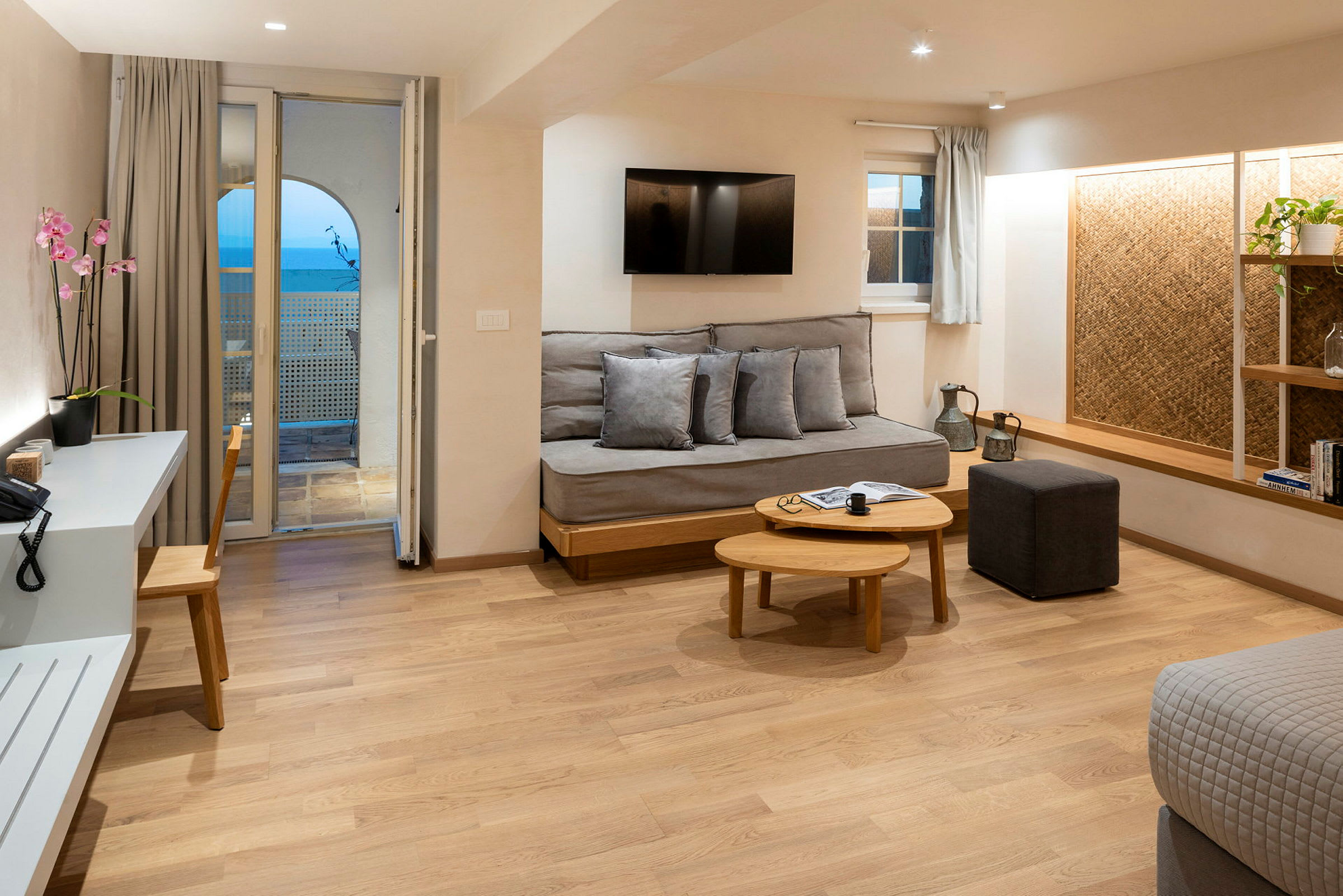 Et moderne hotellrom med tregulv, sofa, salongbord, skrivebord med stol og en balkong med havutsikt. (AI-generert tekst)