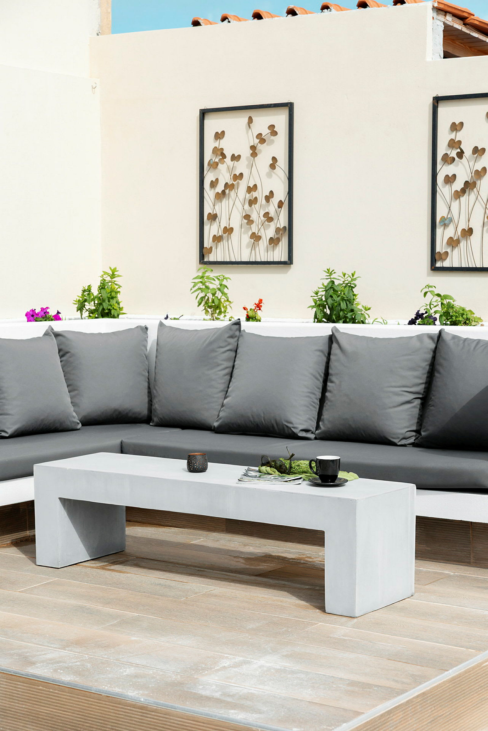 Grå utendørs sofa med puter ved siden av et hvitt betongbord omgitt av planter på en terrasse. (AI-generert tekst)