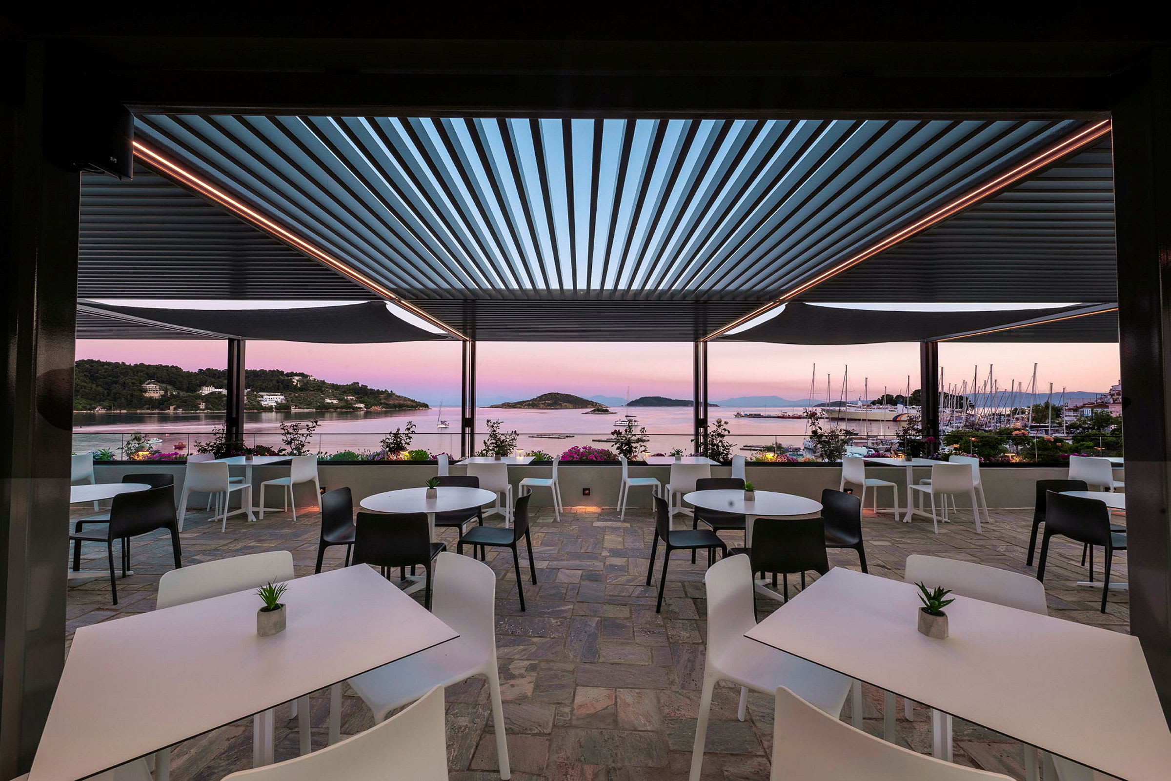 En utendørs terrasse med bord og stoler med utsikt mot havet og seilbåter. (AI-generert tekst)