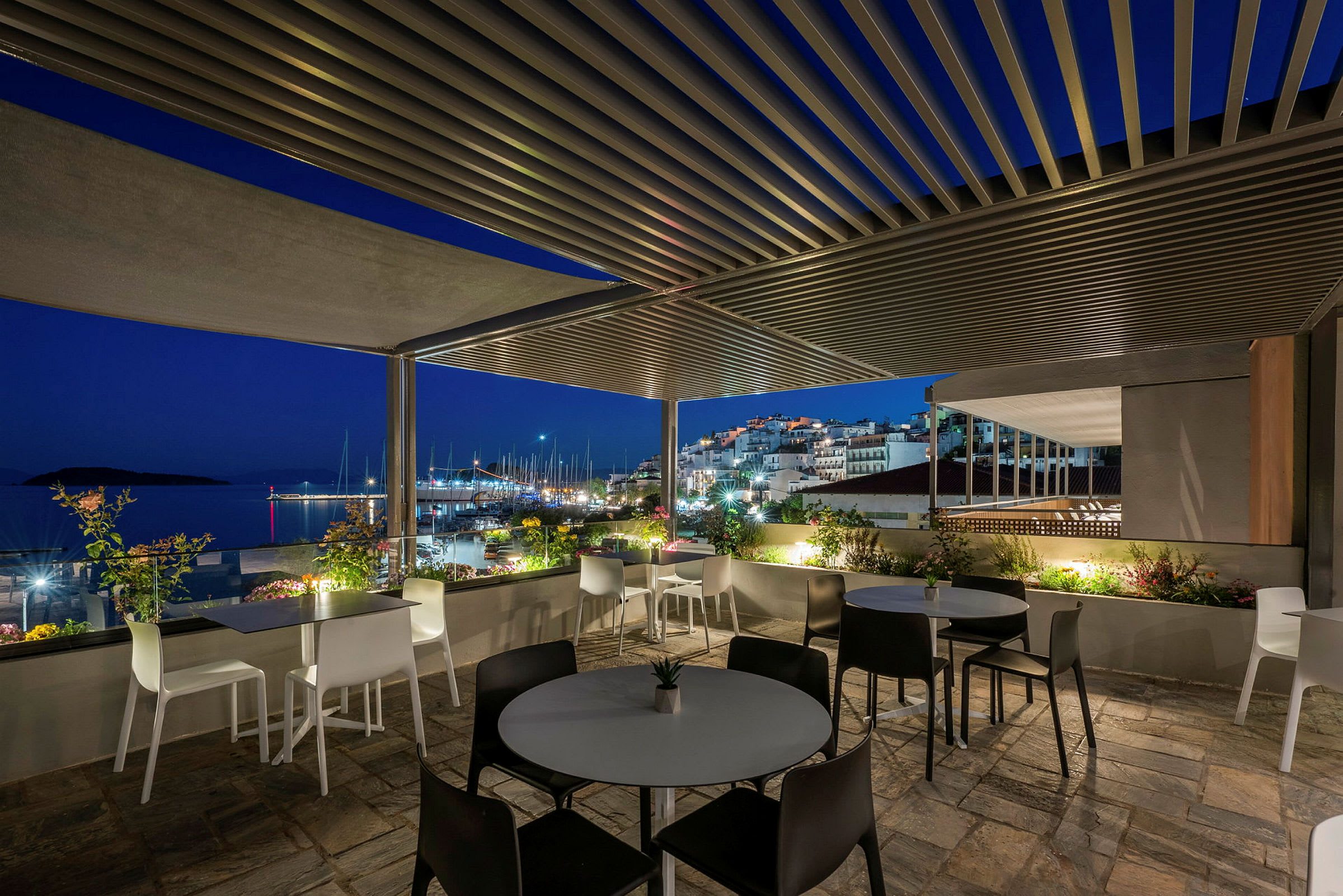 En terrasse med sitteplasser og utsikt over en småbåthavn og bygninger på kveldstid. (AI-generert tekst)
