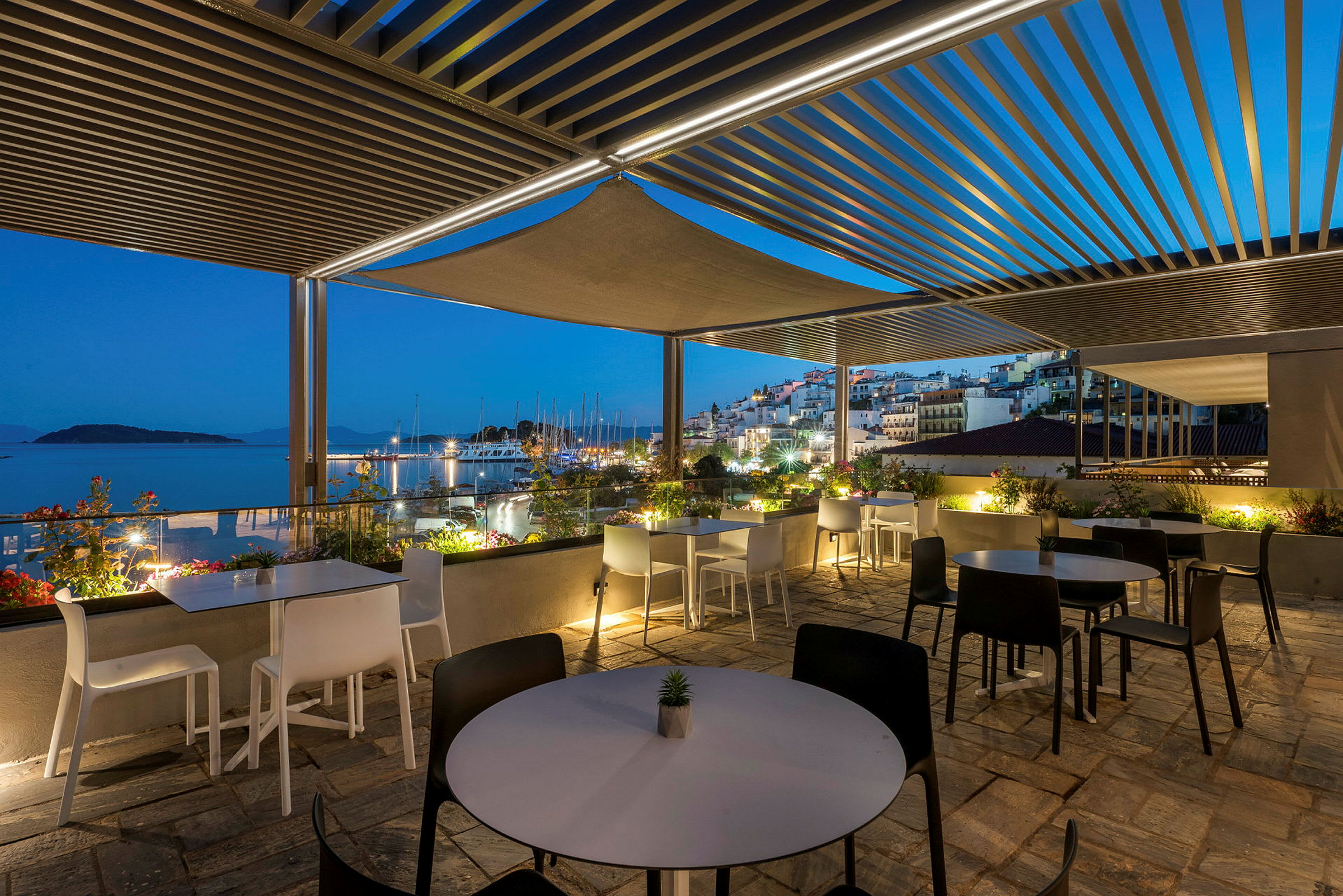 En utendørs terrasse med bord og stoler under en pergola, med utsikt mot en småbåthavn og en opplyst by ved kysten i skumringen. (AI-generert tekst)