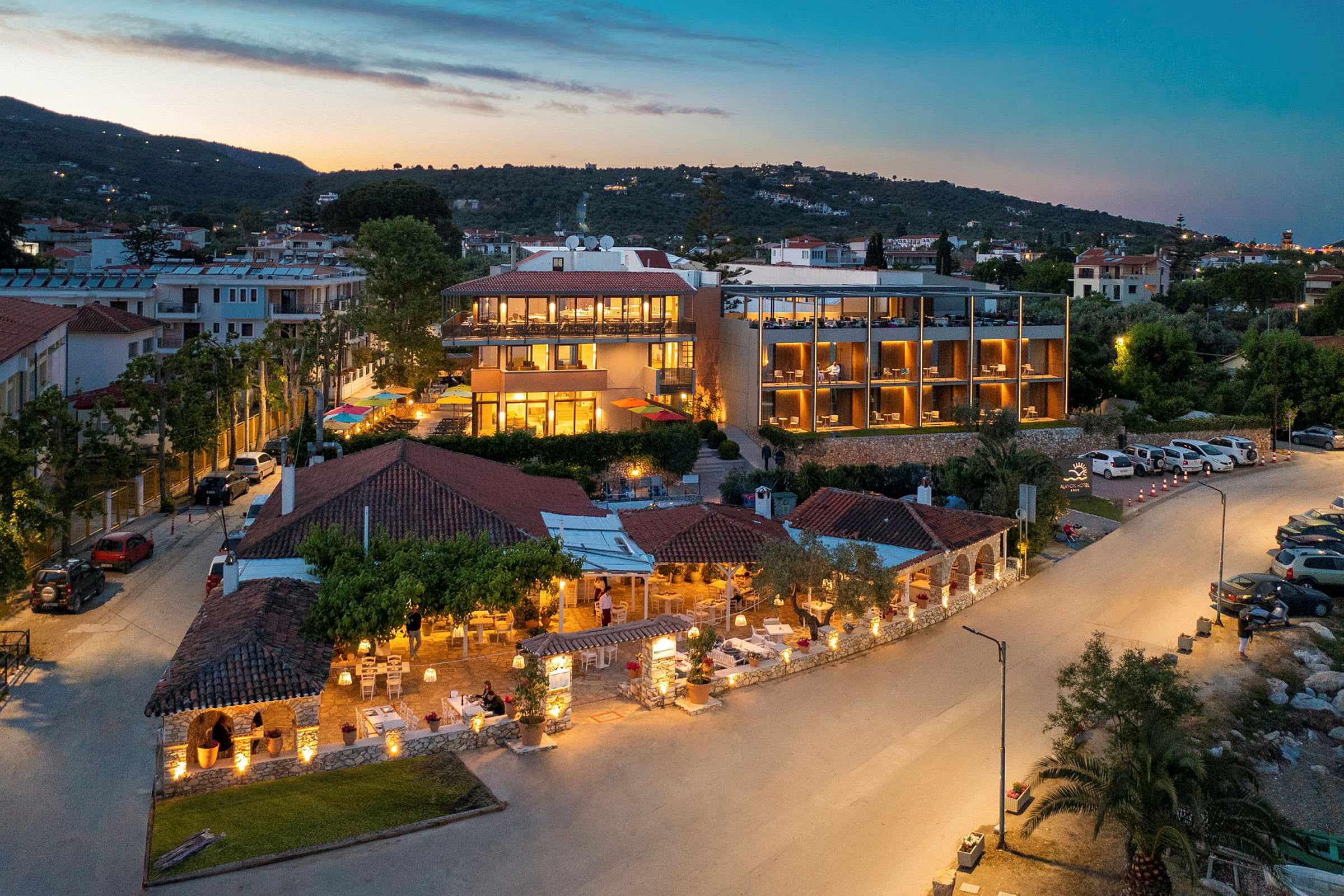 Hotellbygning med kveldsbelysning, omgitt av grøntområder og parkeringsplasser. (AI-generert tekst)