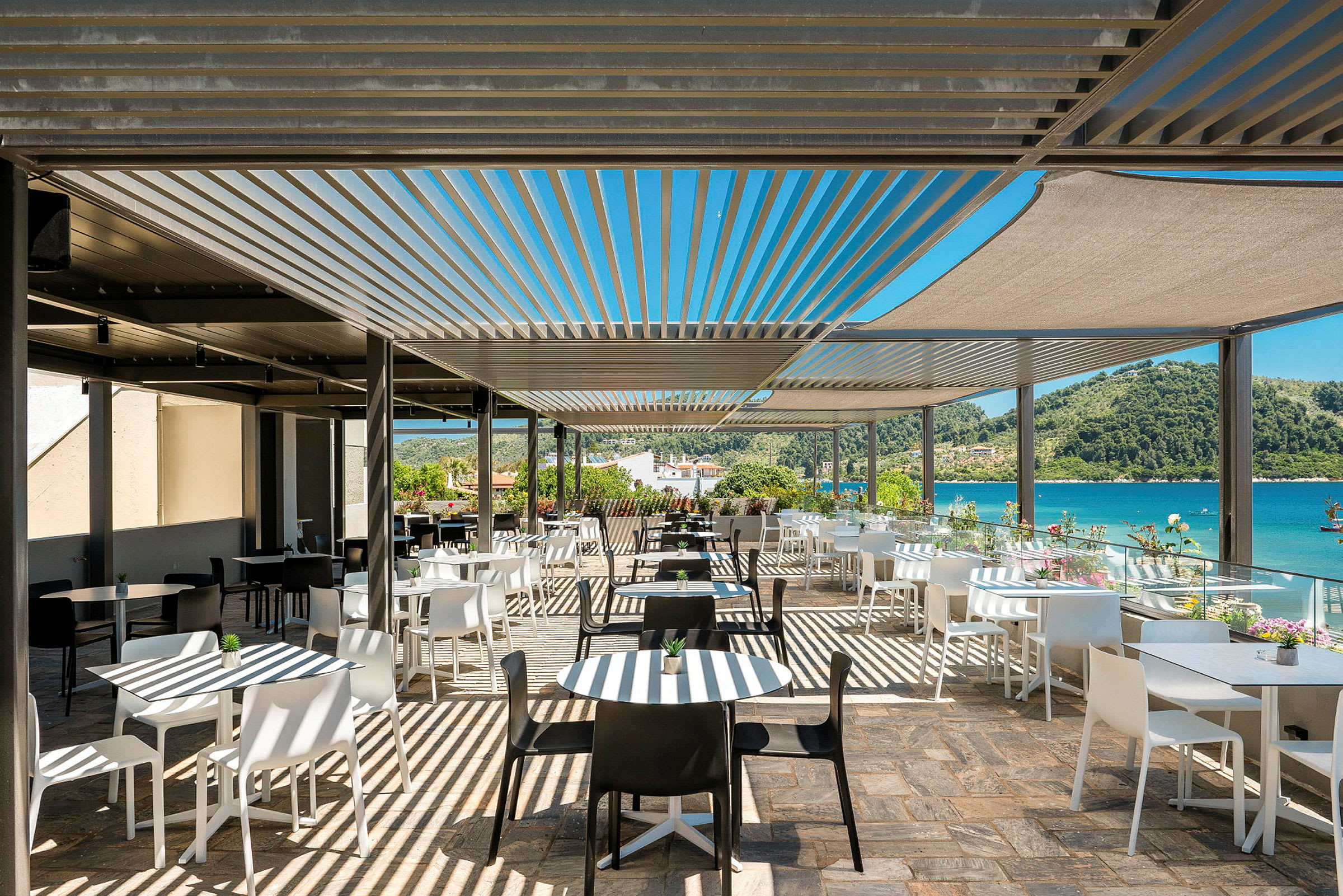 En utendørsrestaurant med hvite og svarte stoler og bord under solskjerming, med utsikt over havet og omkringliggende grøntområder. (AI-generert tekst)