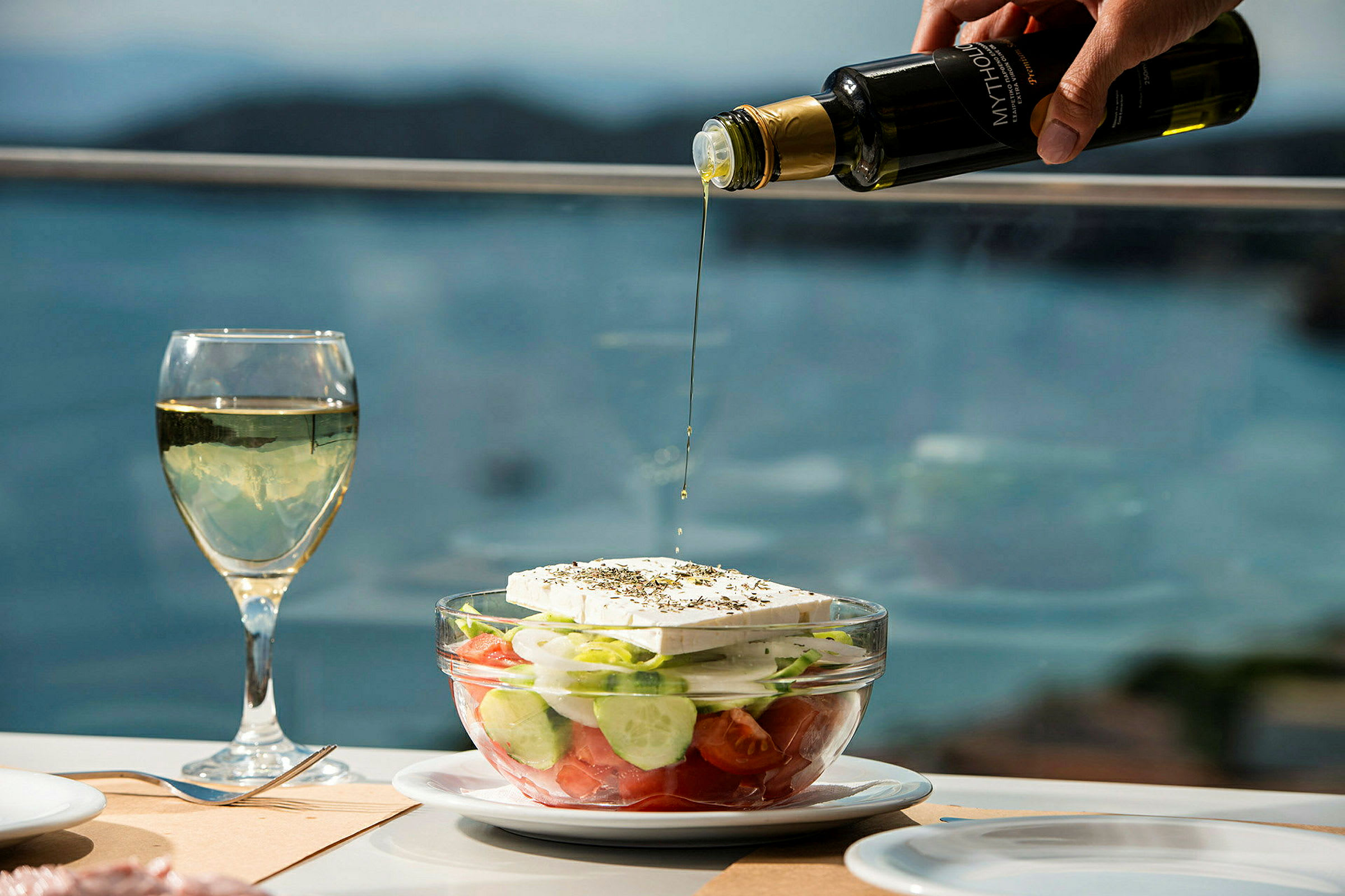En gresk salat i en glasskål ved et dekket bord. En hånd heller olivenolje over salaten. (AI-generert tekst)