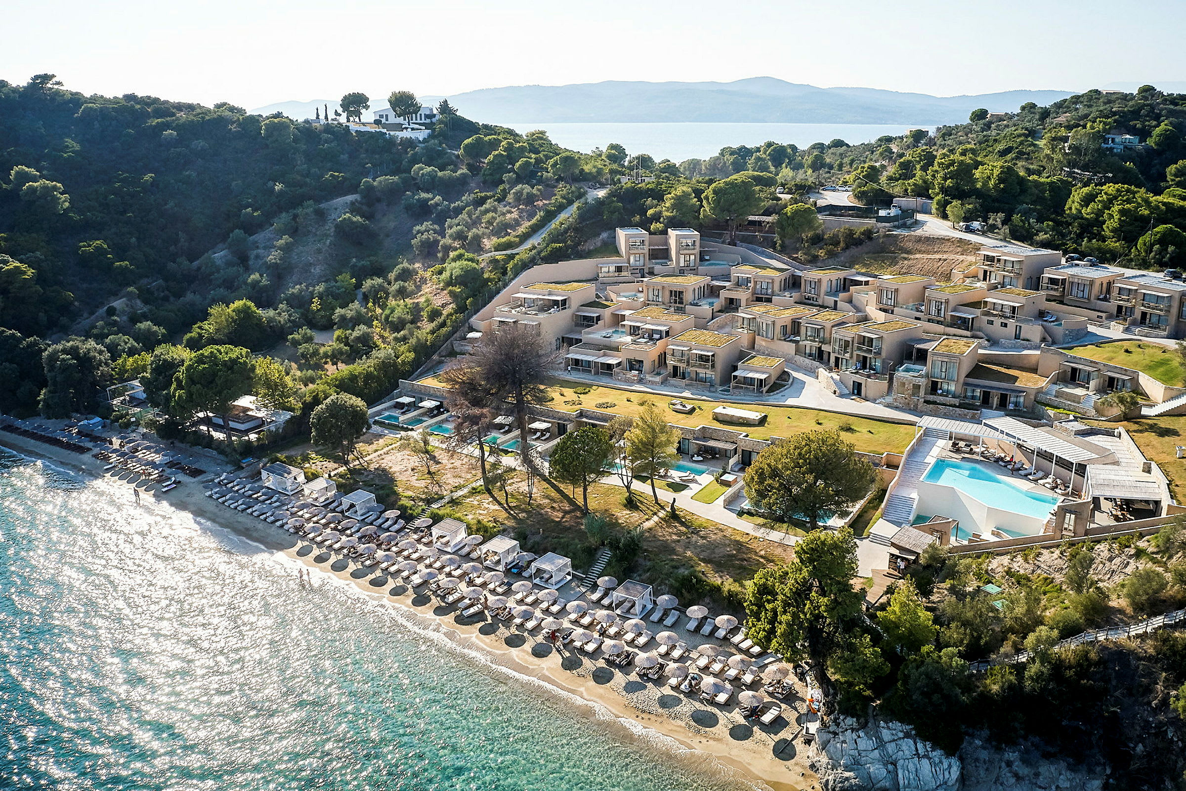 Flyfoto av resort med basseng, strand med parasoller og solsenger, samt omkringliggende vegetasjon. (AI-generert tekst)