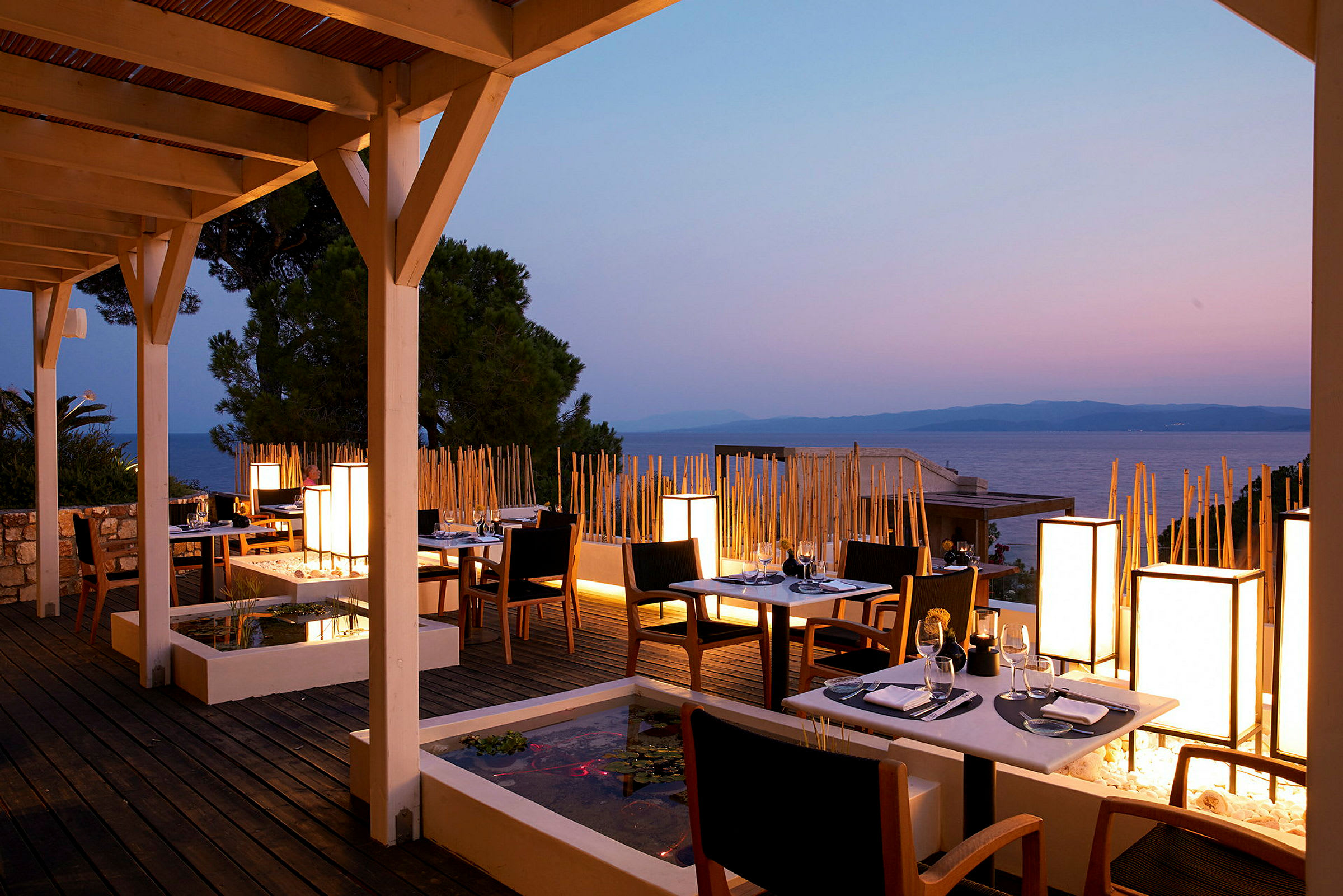 En restaurantterrasse med bord og stoler ved solnedgang, havet i bakgrunnen. (AI-generert tekst)