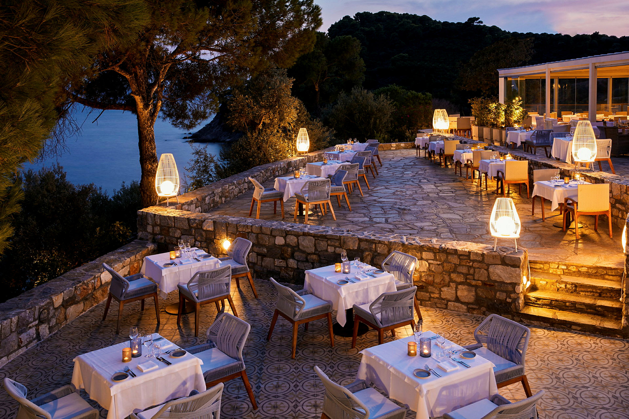 En restaurantterrasse med belysning, dekkede bord og utsikt over havet ved solnedgang. (AI-generert tekst)