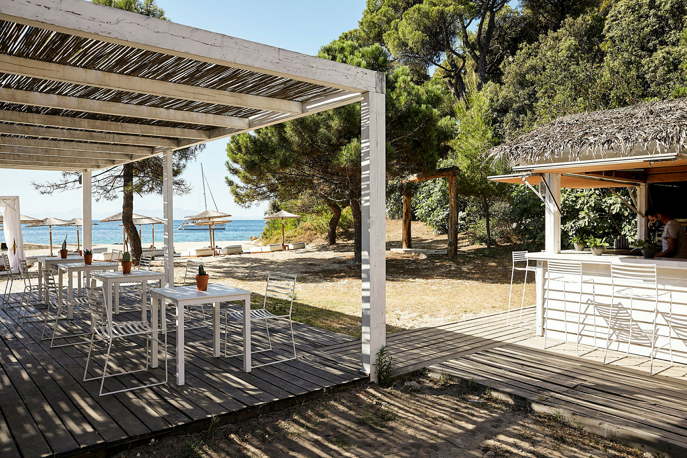 En strandbar med sitteplasser under en trepergola, nærliggende parasoller og en skog i bakgrunnen. (AI-generert tekst)