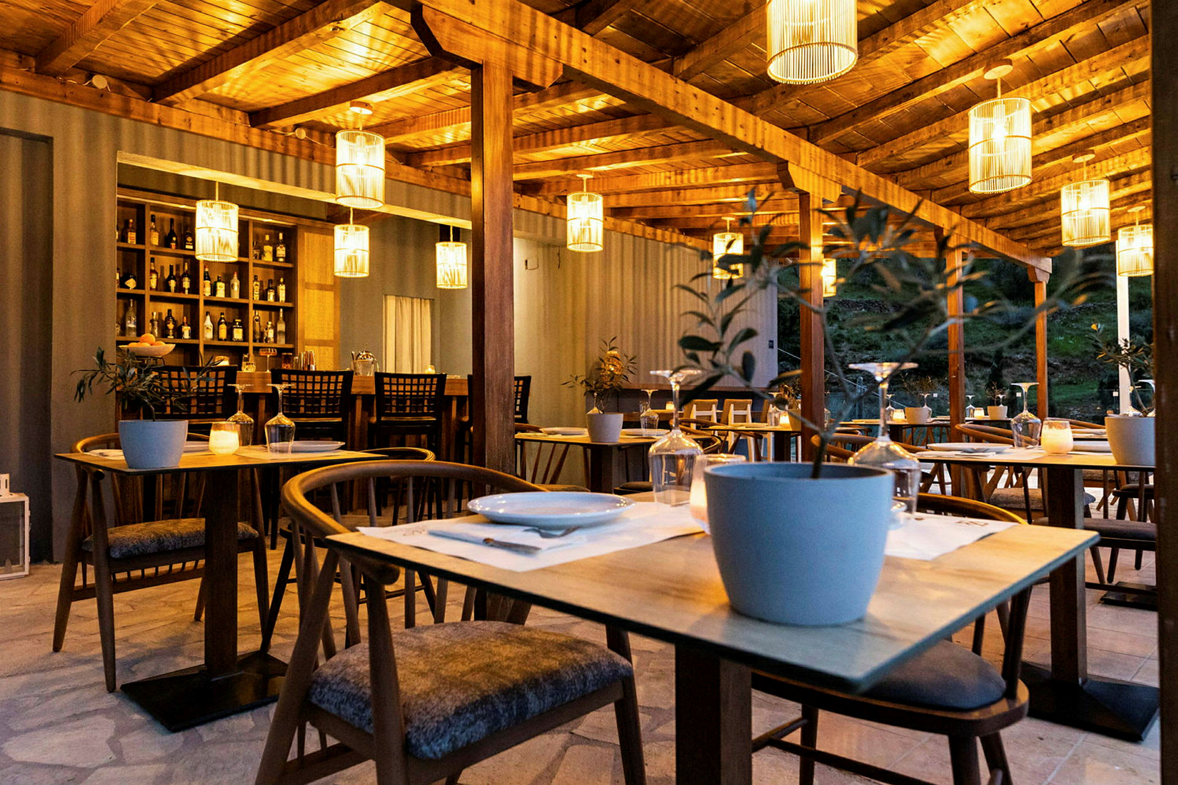 En utendørs restaurant med trebord og stoler, dekorert med hengende lampeskjermer og potteplanter. (AI-generert tekst)