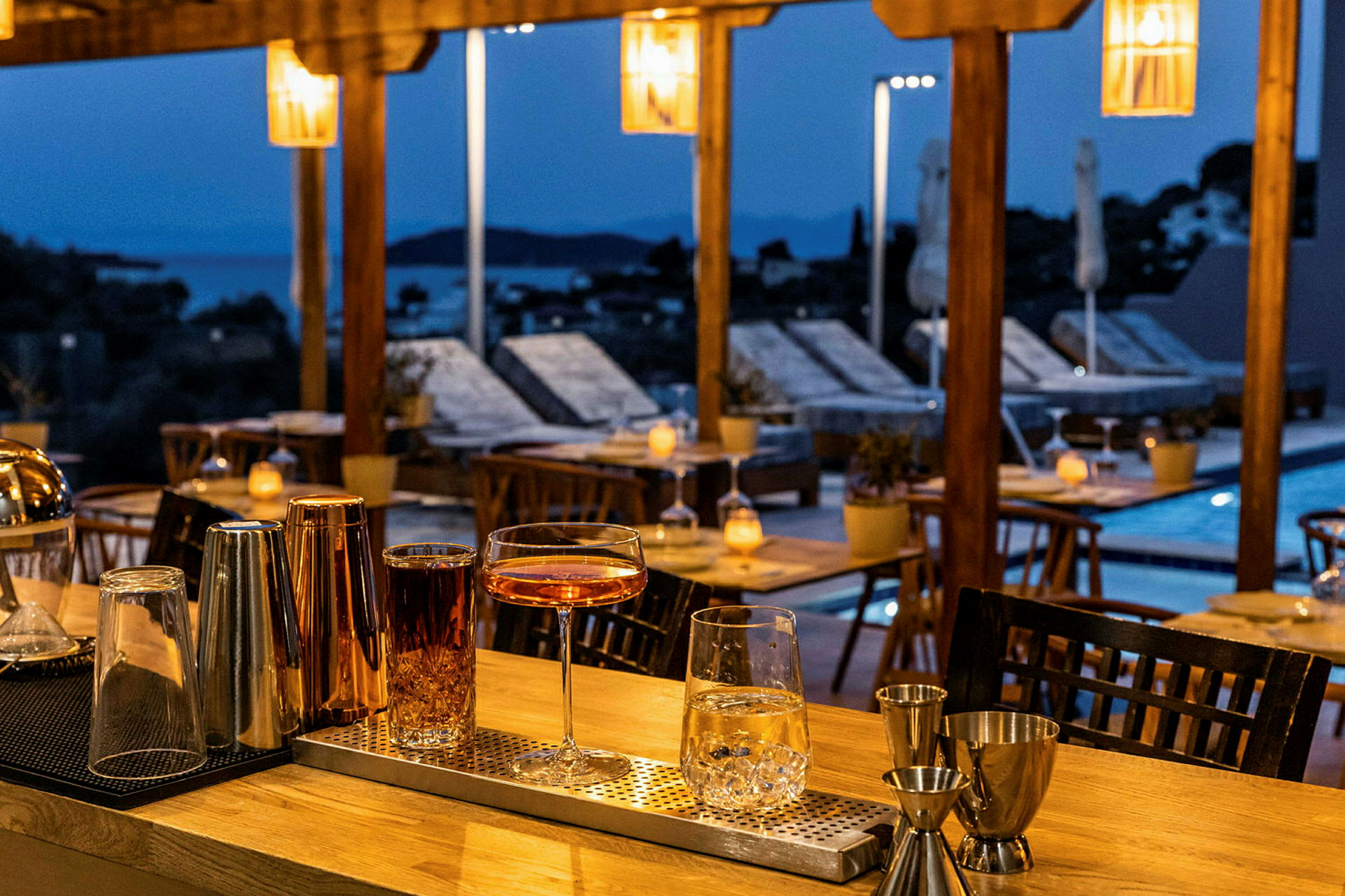 Cocktailglass og drinker står på en bardisk ved en kveldsbelyst terrasse. (AI-generert tekst)