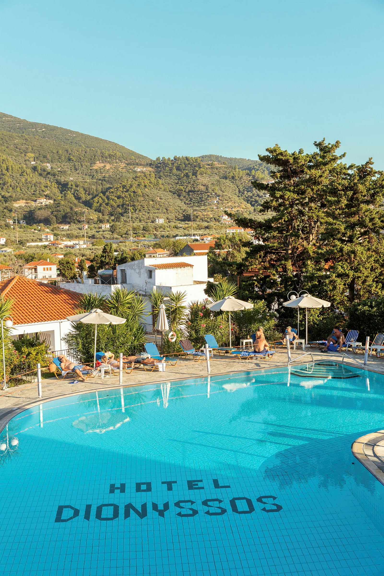 Bilde av et basseng ved Hotel Dionyssos med solsenger og parasoller rundt. I bakgrunnen ses trær og et fjellandskap. (AI-generert tekst)