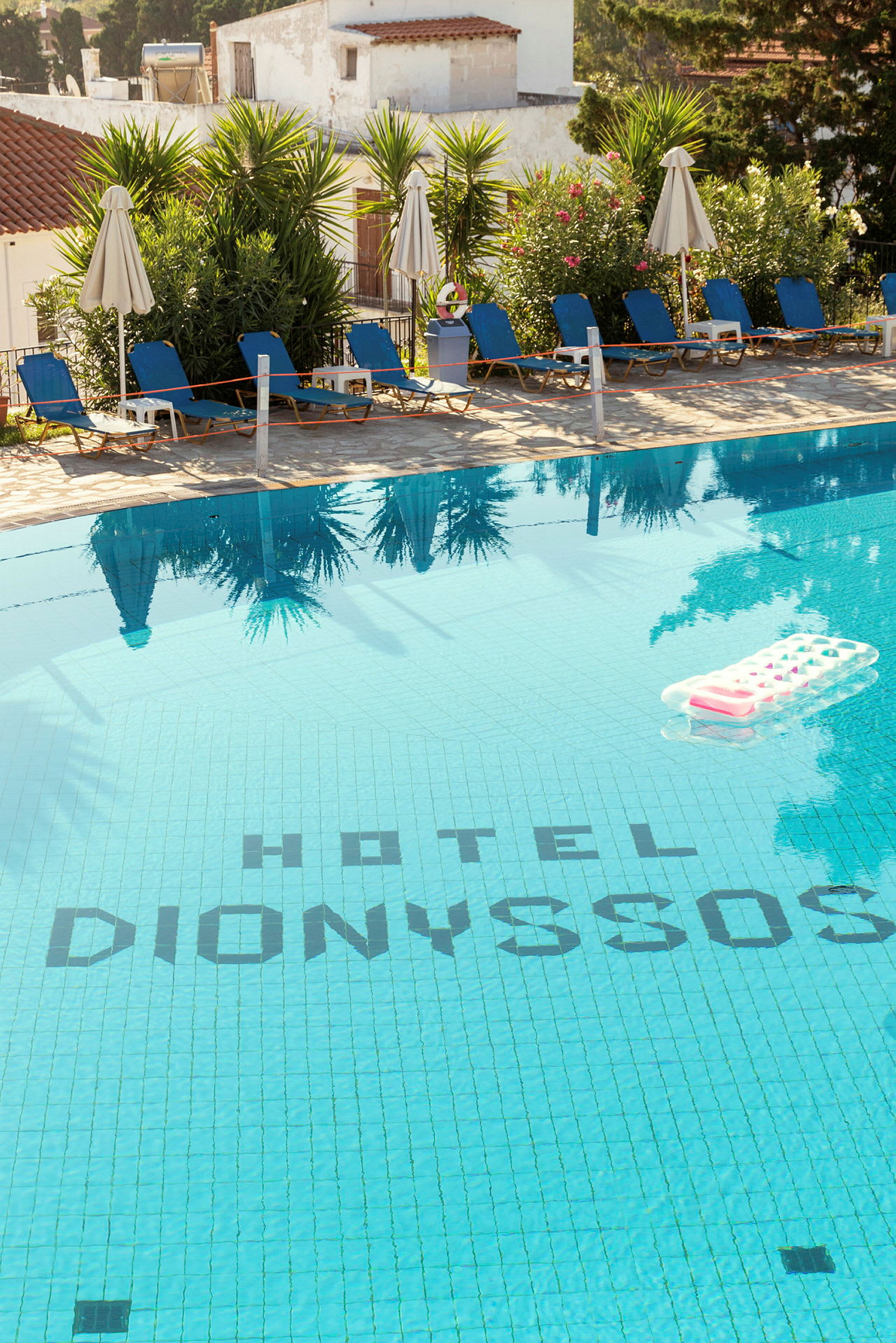 Utendørsbasseng ved Hotel Dionysos med solsenger og parasoller omgitt av grøntområder. (AI-generert tekst)