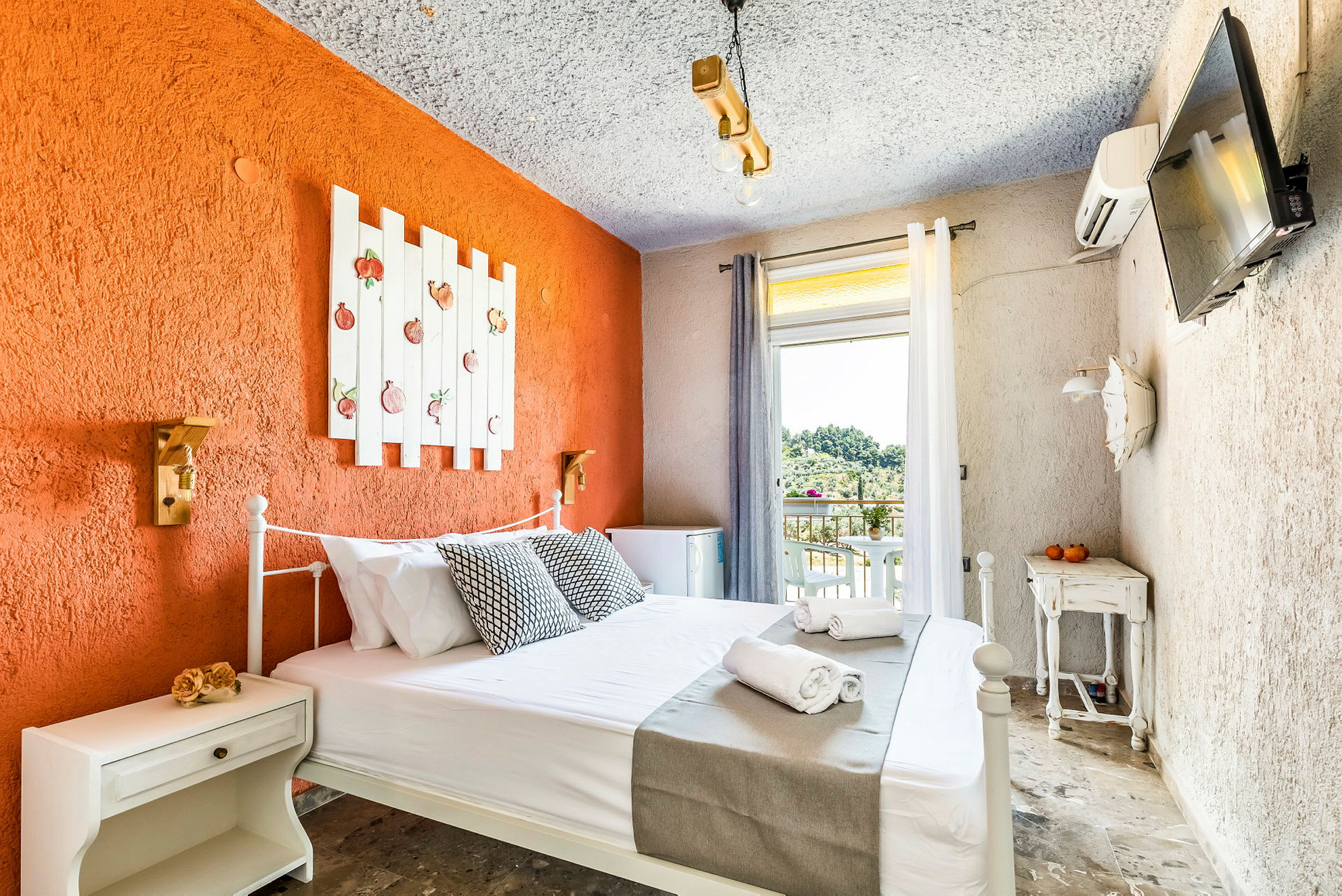 Hotellrom med en oransje aksentvegg, dekorert med en hvit treramme med fruktmotiv, en seng med hvite laken og grå sengeteppe, og en liten balkong med utsikt mot grøntområder. (AI-generert tekst)