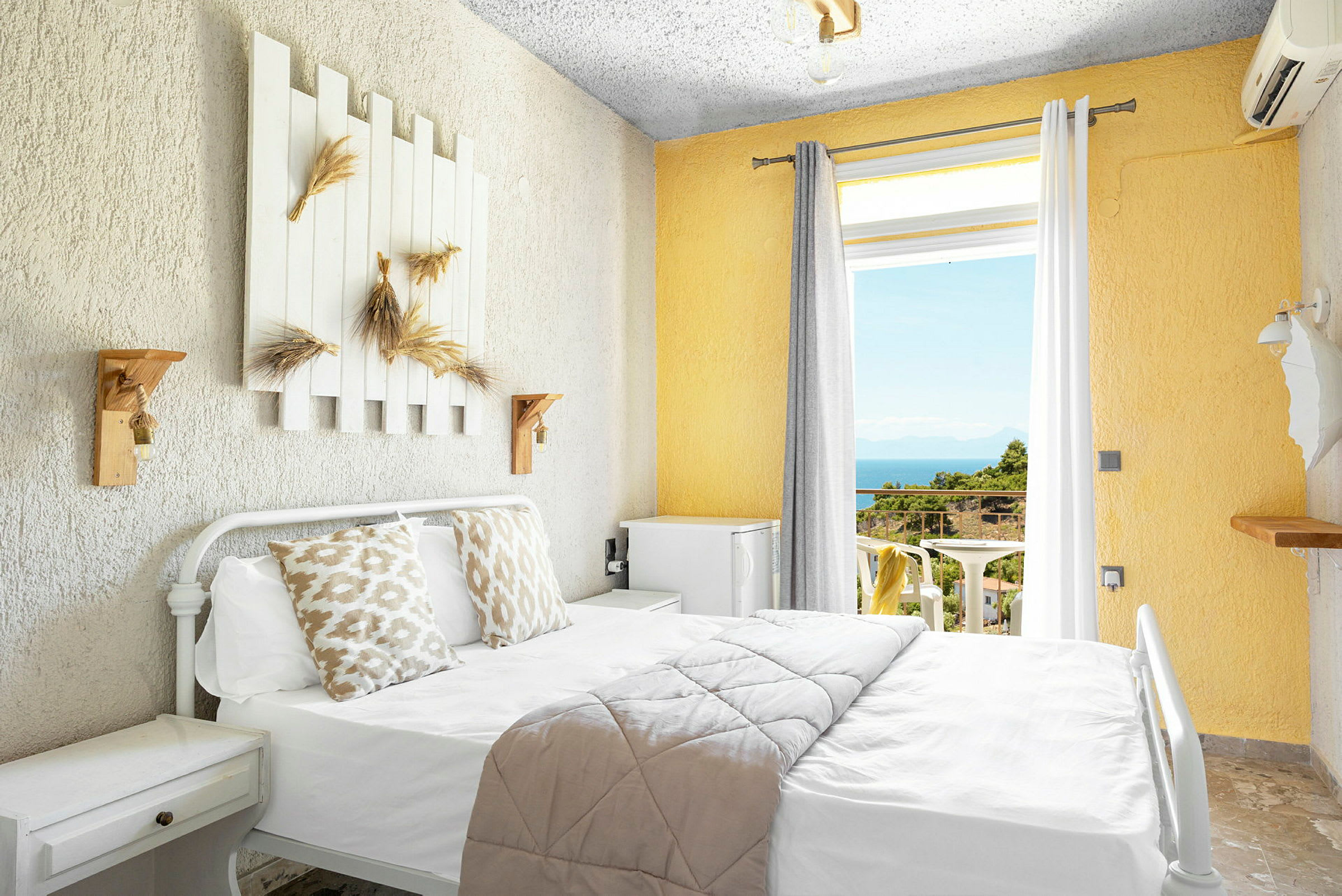Lyst hotellrom med seng, dekorerte puter og utsikt mot balkong med hav og grøntområder. (AI-generert tekst)