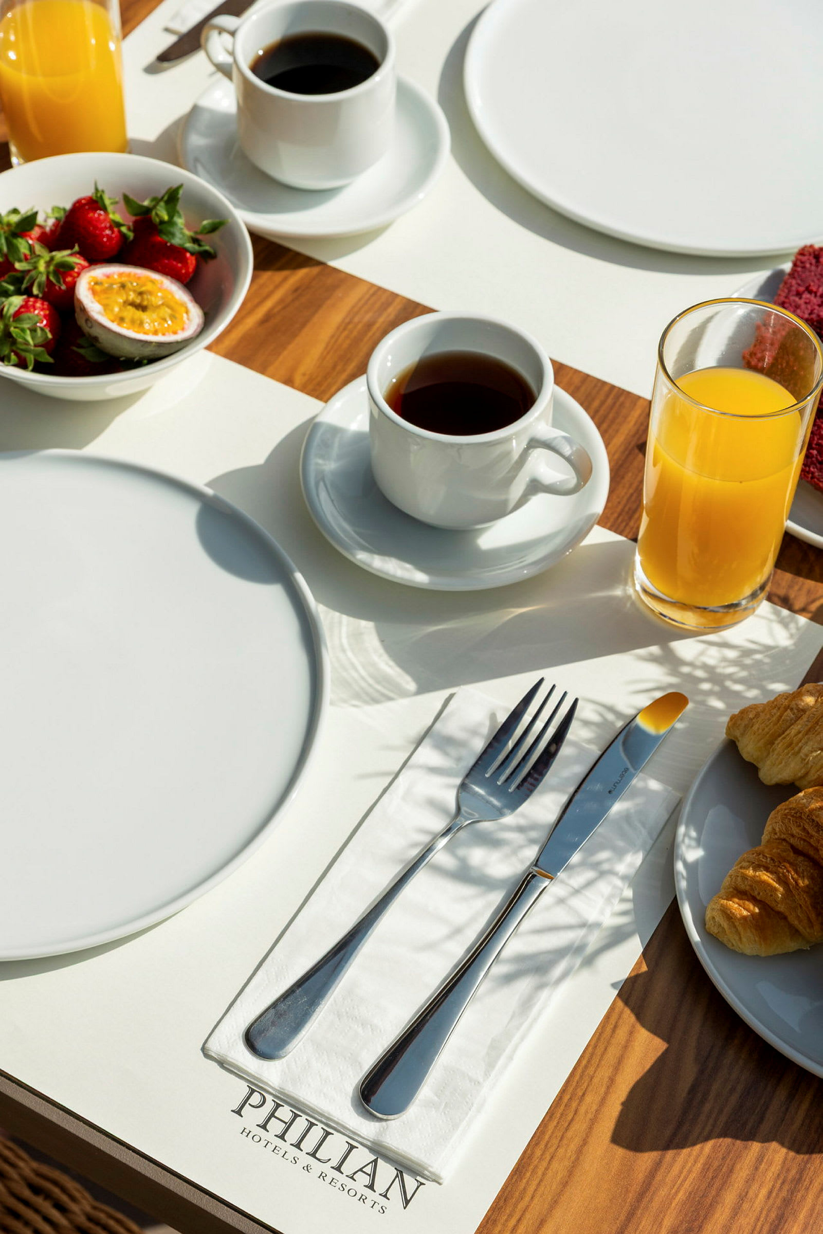 Frokostbord med tallerkener, bestikk, kaffe, juice, jordbær og croissanter på en treoverflate. (AI-generert tekst)