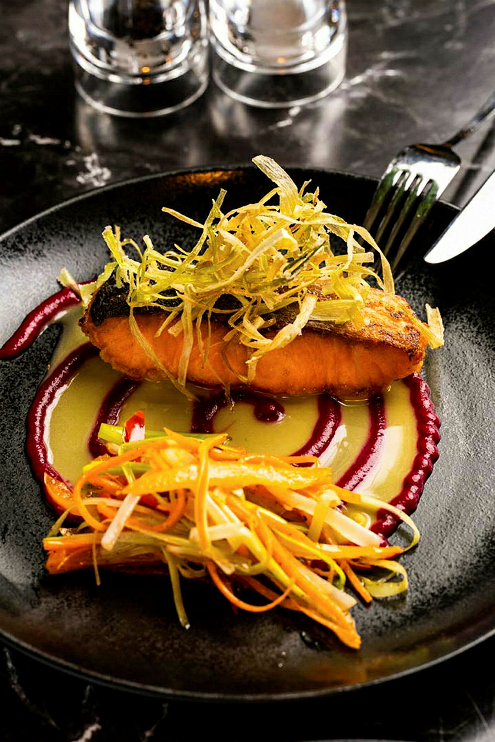En tallerken med en rett bestående av tilberedt fisk, topping av grønnsaker og saus. Bestikk og glass er synlige i bakgrunnen. (AI-generert tekst)