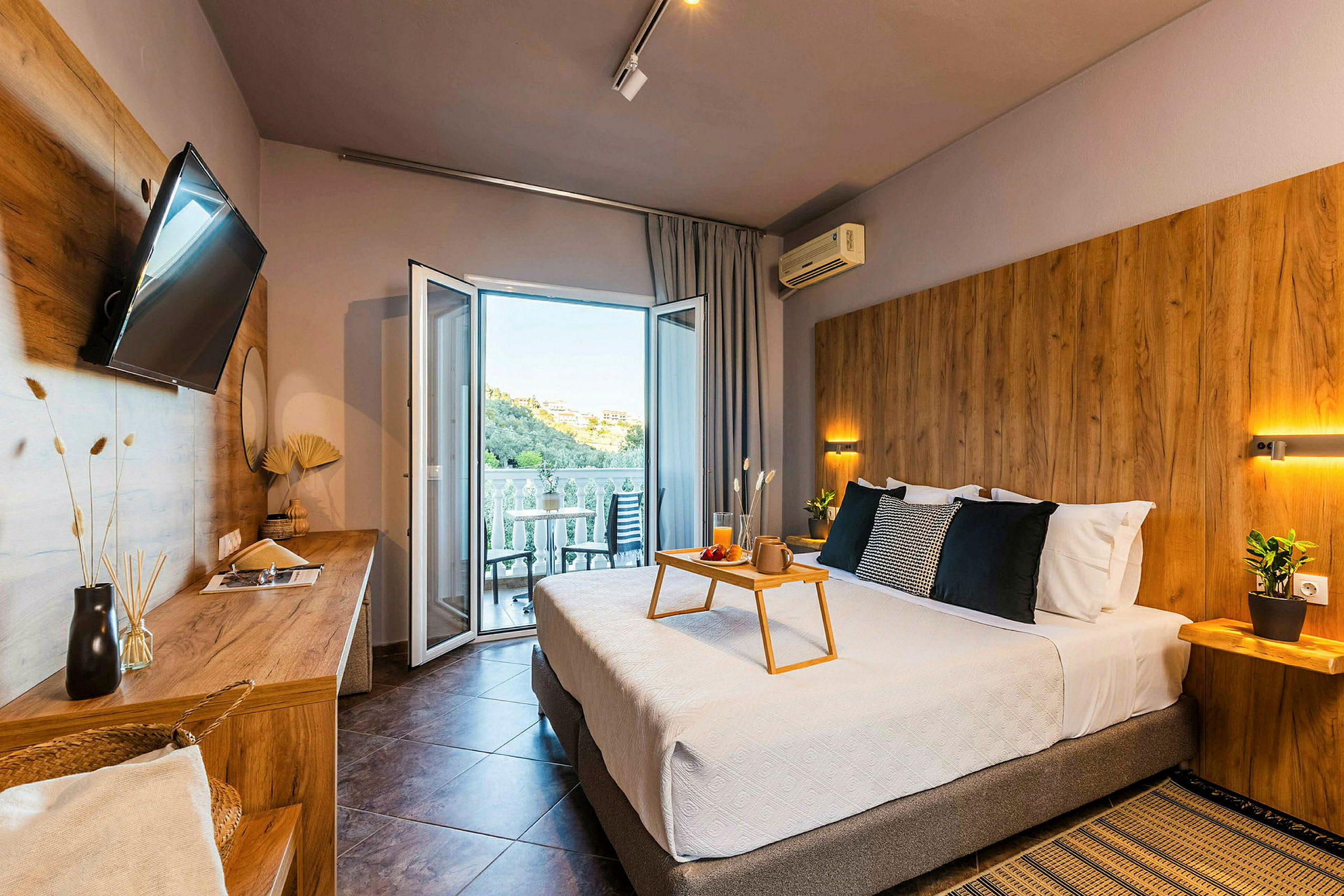 Moderne hotellrom med dobbeltseng, sittemøbler på balkongen og utsikt over en grønn ås. (AI-generert tekst)