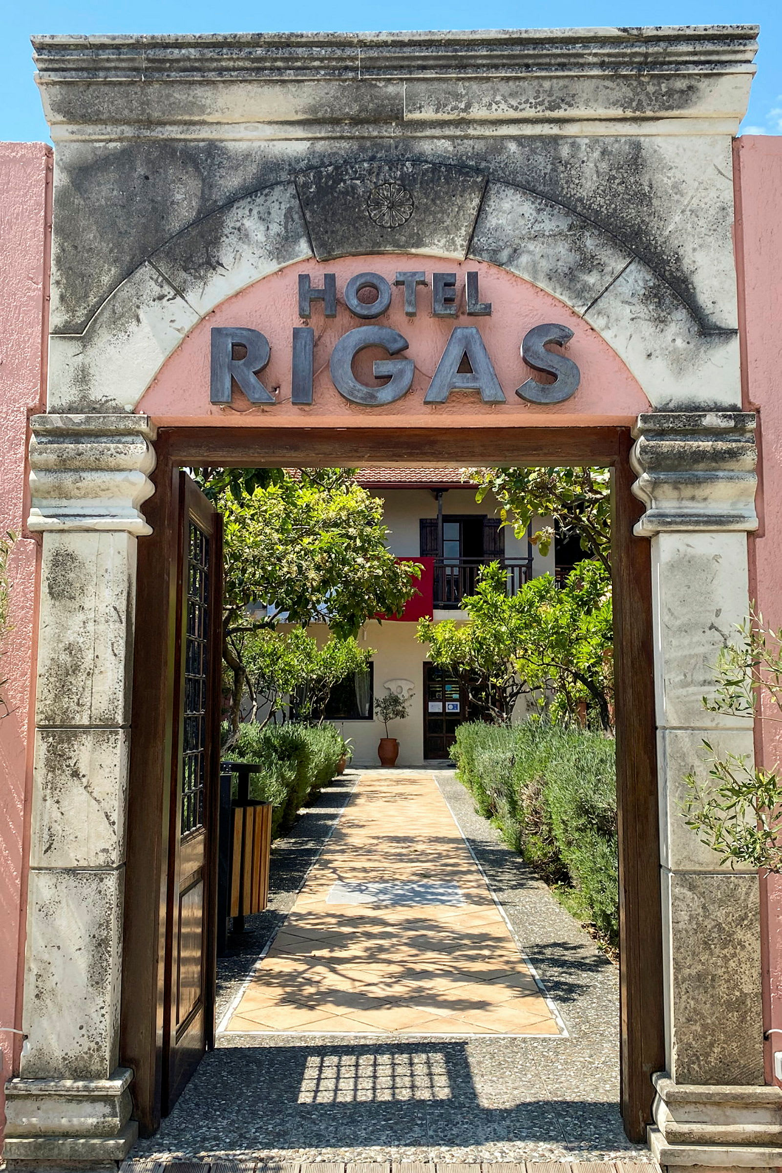Bilde av en hotellinngang med teksten 'Hotel Rigas', omgitt av en hage og en sti mot døren. (AI-generert tekst)