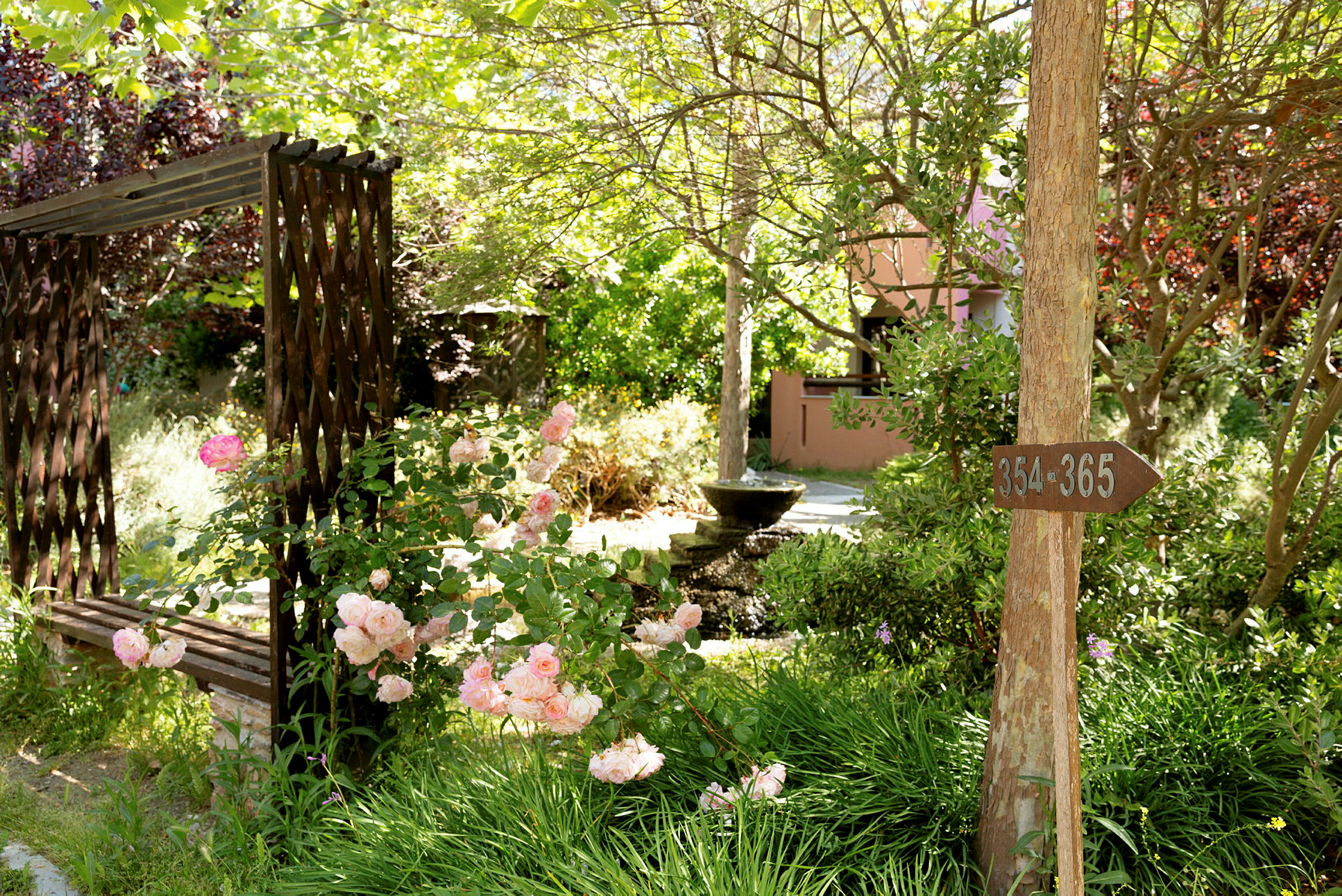 En hage med en trebenk, blomster og en fontene. Et treskilt viser numrene 354-365. (AI-generert tekst)