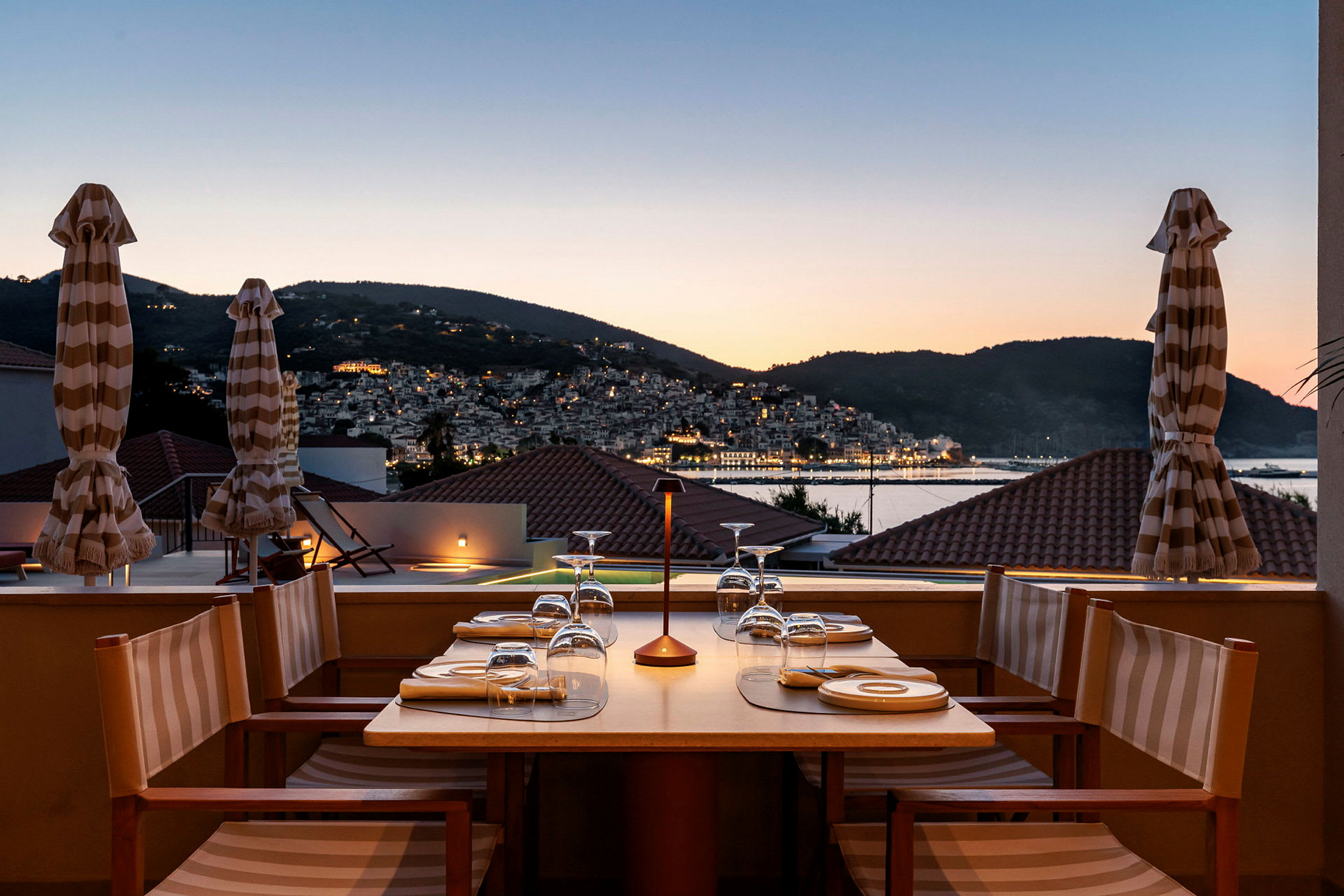 Bord dekket til middag på en terrasse med utsikt over en by og fjell i skumring. (AI-generert tekst)