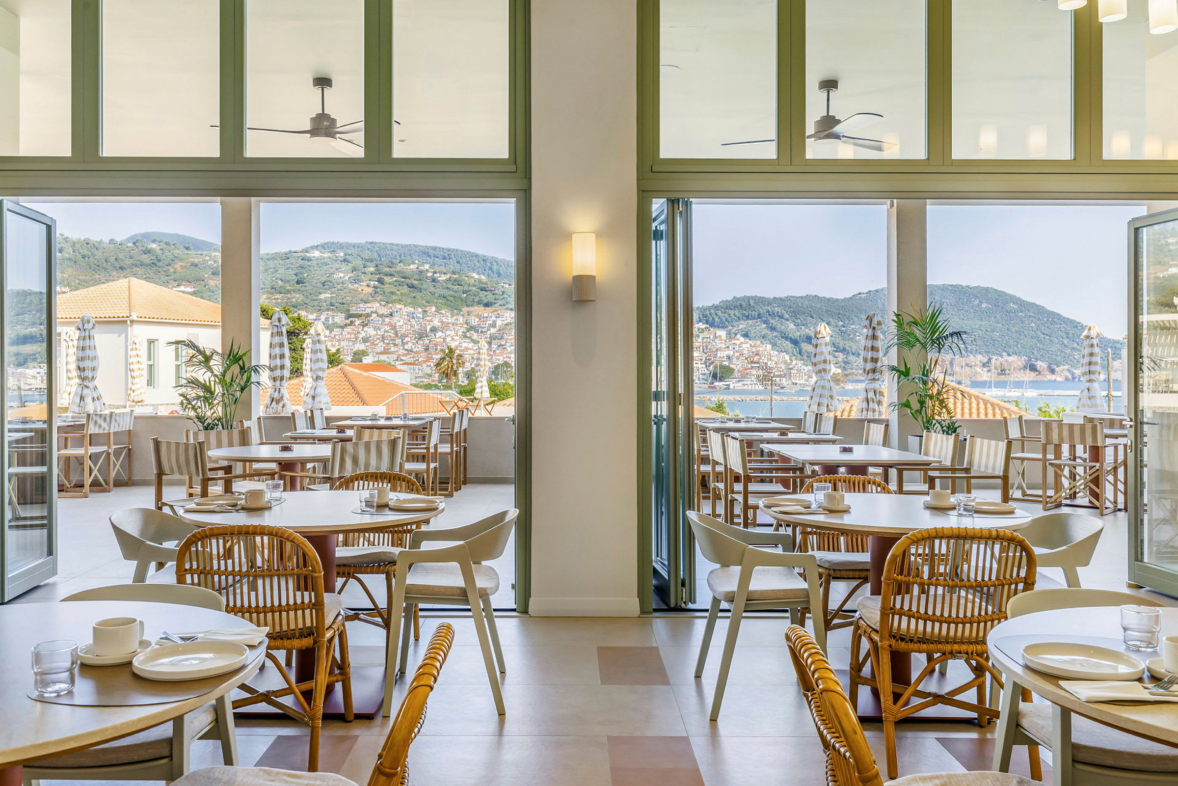 Bilde av en lys og luftig restaurantområde med bord og stoler innendørs samt en terrasse med utsikt over havet og byen. (AI-generert tekst)