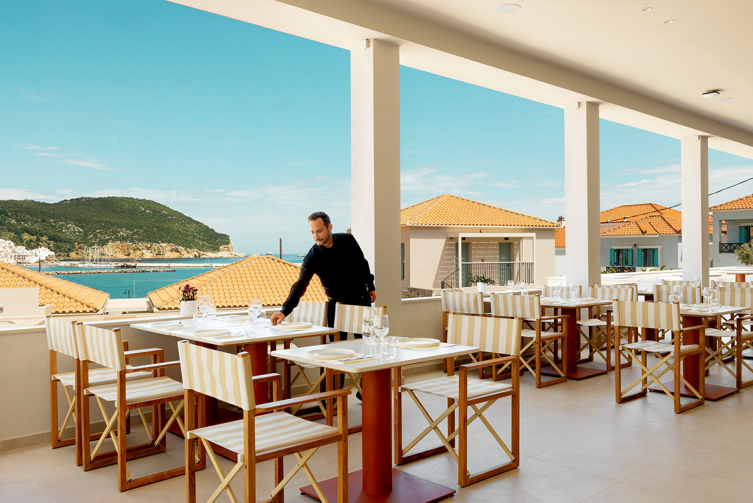 Restaurant med bordoppsett, utsikt over havet og terrasse, med bakgrunn av terrakottatak og en frodig åsside. (AI-generert tekst)