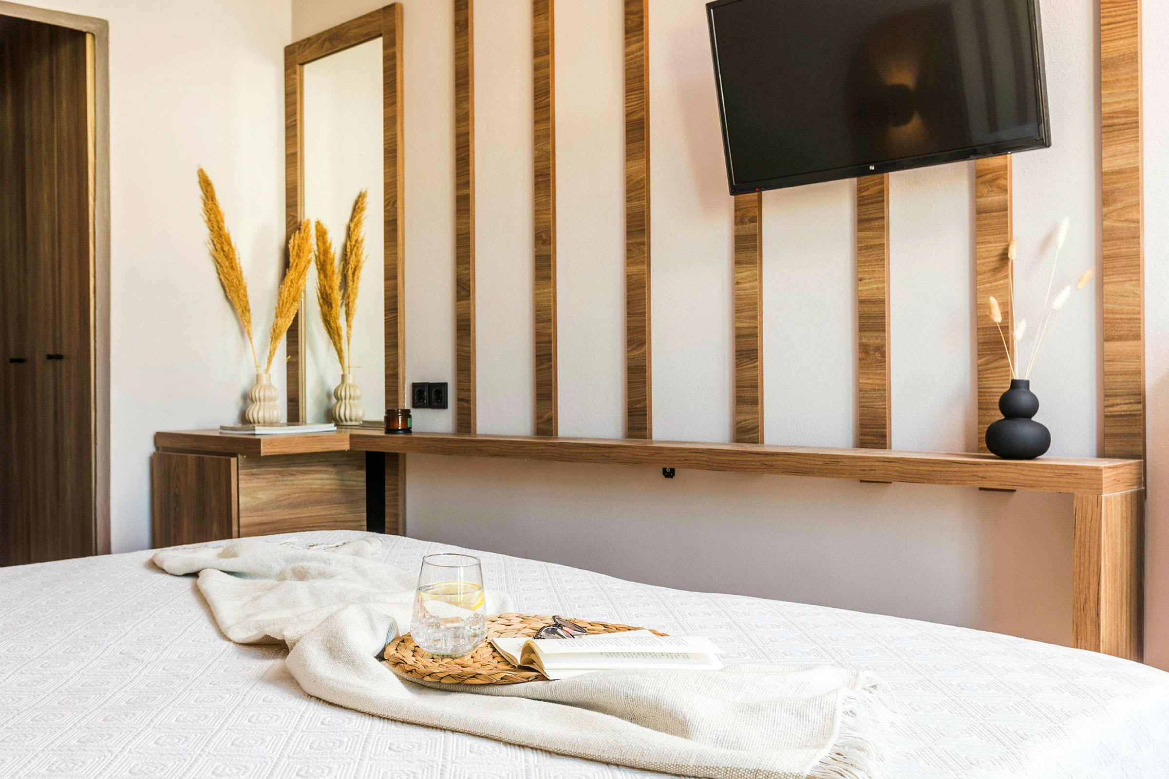 Et moderne designet hotellrom med et hvitt sengeteppe, et glass vann og en bok på et trebord, samt en vegg med trepaneler, et speil og en TV. (AI-generert tekst)
