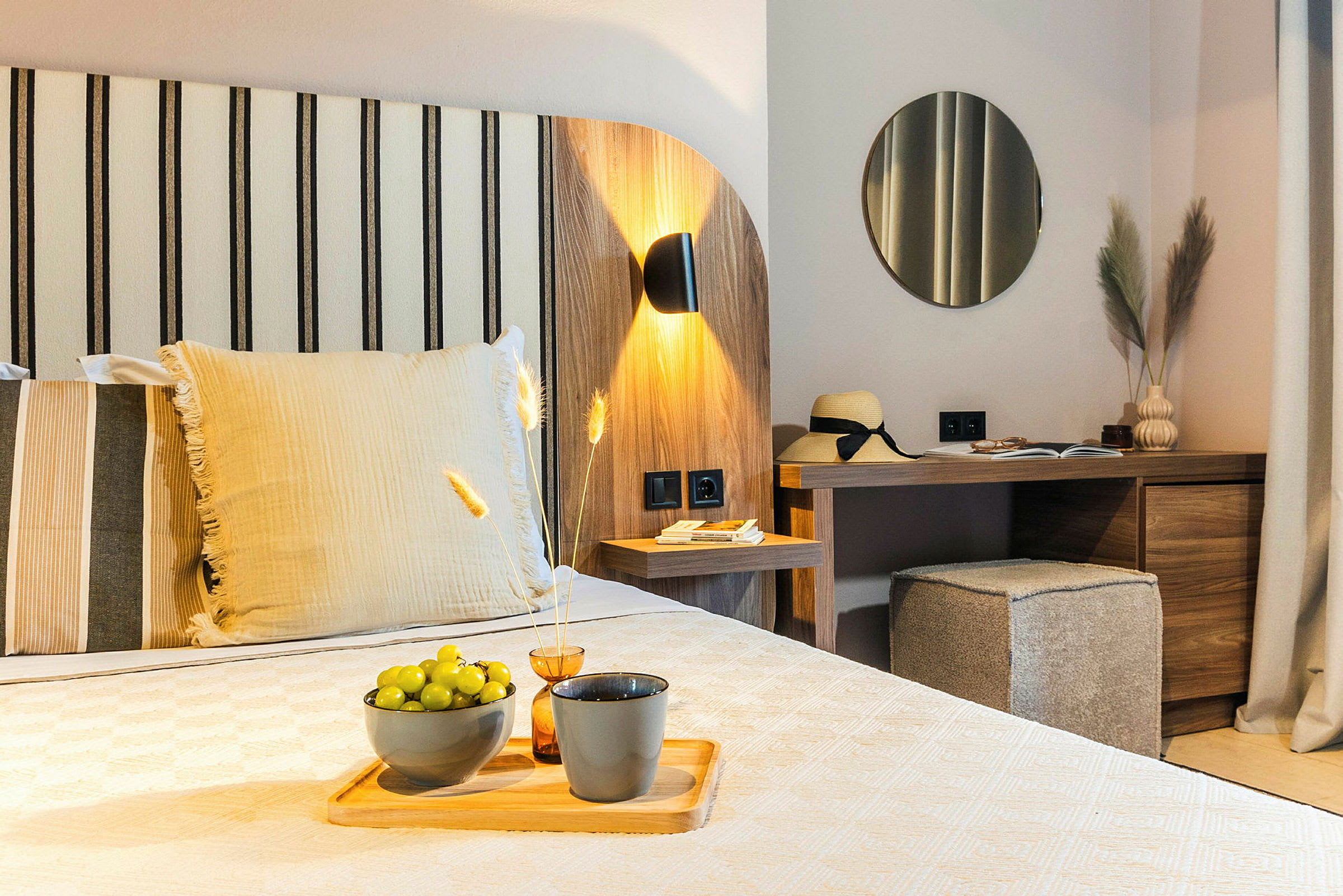 Et moderne hotellrom med tredekor, en seng med dekorative puter, et skrivebord med speil og dekorative gjenstander, og grønne druer på et fat. (AI-generert tekst)