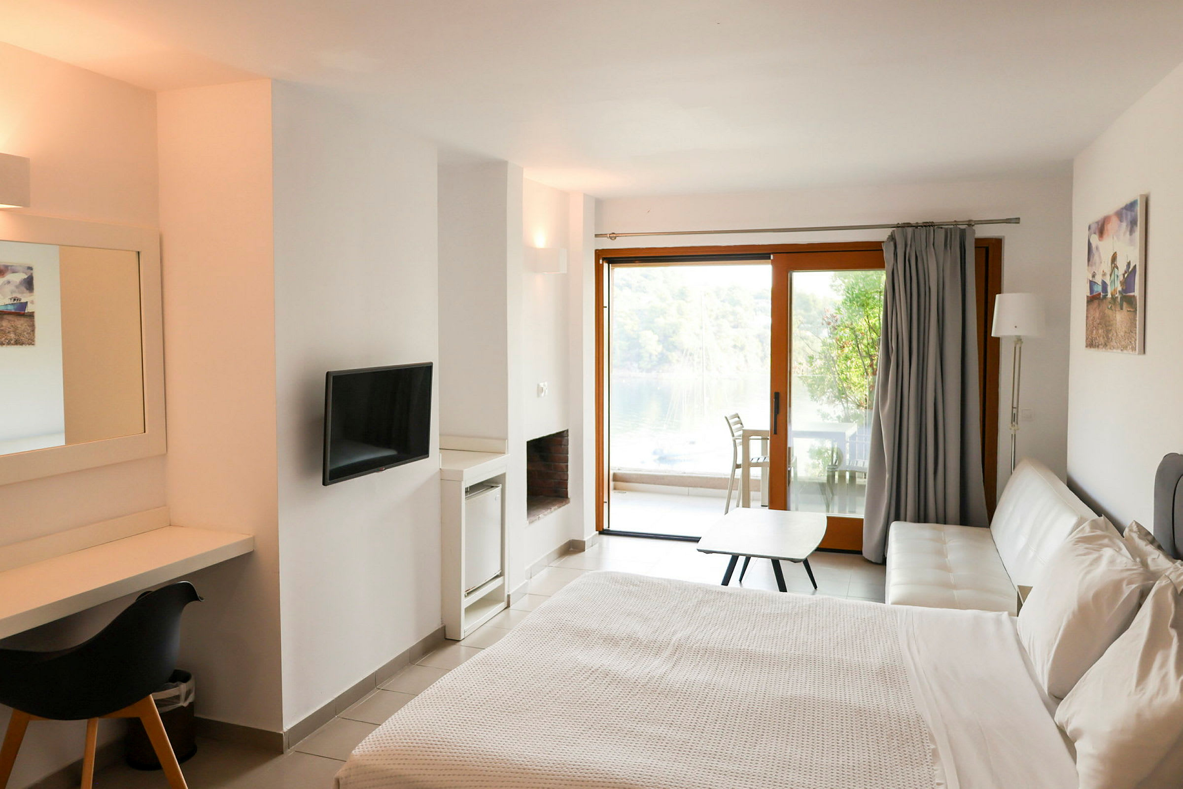 Et moderne hotellrom med en dobbeltseng, skrivebord, TV og en hvit sofa. I bakgrunnen er det en glassdør som fører til en møblert terrasse med utsikt mot natur og vann. (AI-generert tekst)
